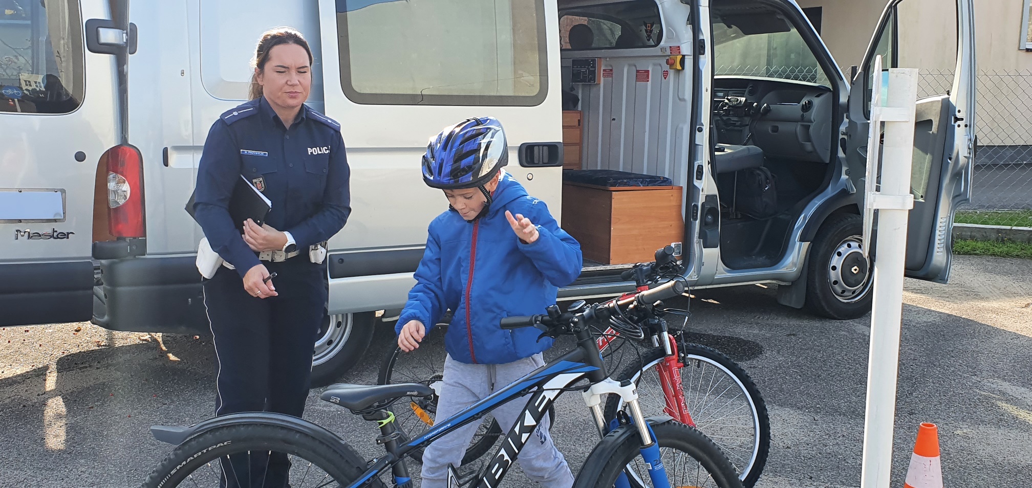 Dziecko w niebieskim kasku stoi za rowerem. Po jego praqwej stronie stoi polcjantka