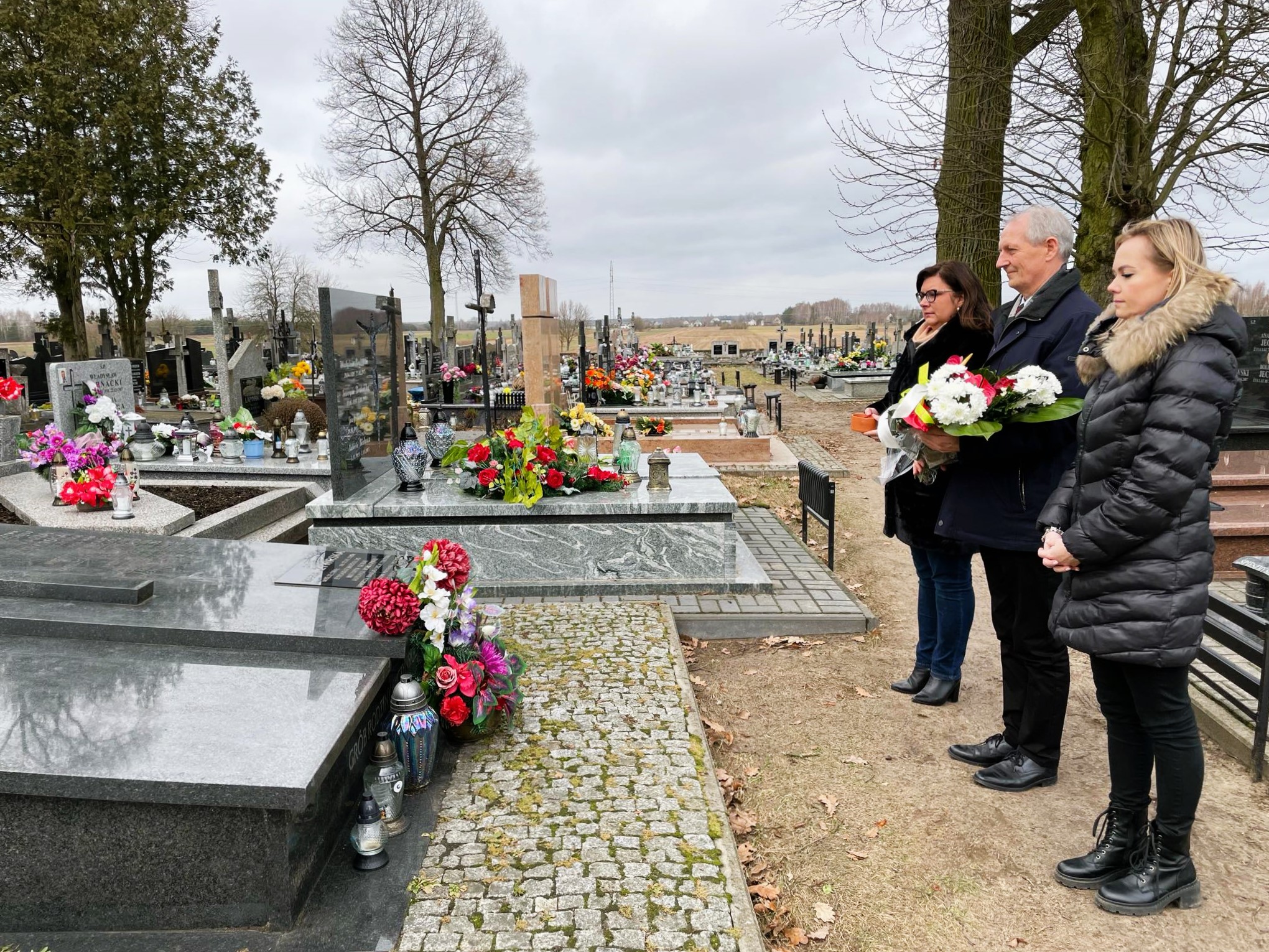 Cmentarz parafialny w Brańszczyku, delegacja samorządowców (wójt i dwie kobiety) podczas złożenia kwiatów i zniczy na grobie Marii Żywirskiej.
