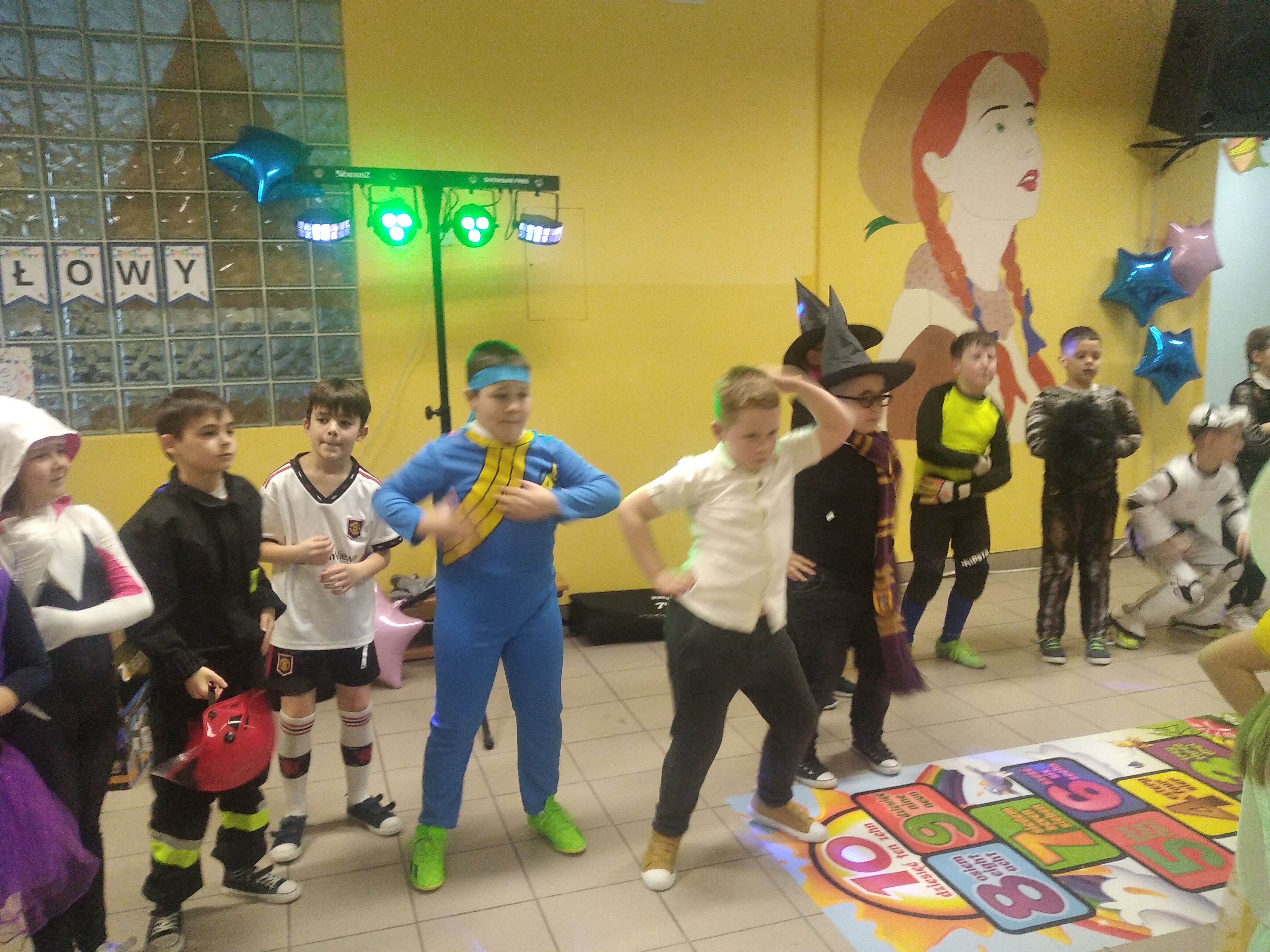 Chłopcy przebrani w niebieski strój Ninja, w piłkarza i strażaka oraz chłopiec w białej koszuli tańczą w rytm muzyki. Po prawej stronie zdjęcia widać Harrego Pottera, rycerza oraz kolejnego piłkarza-bramkarza.