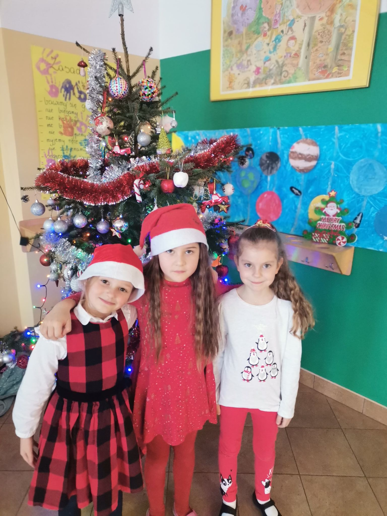 Trzy dziewczynki z czapkach św. Mikołaja stoją przed udekorowaną choinką