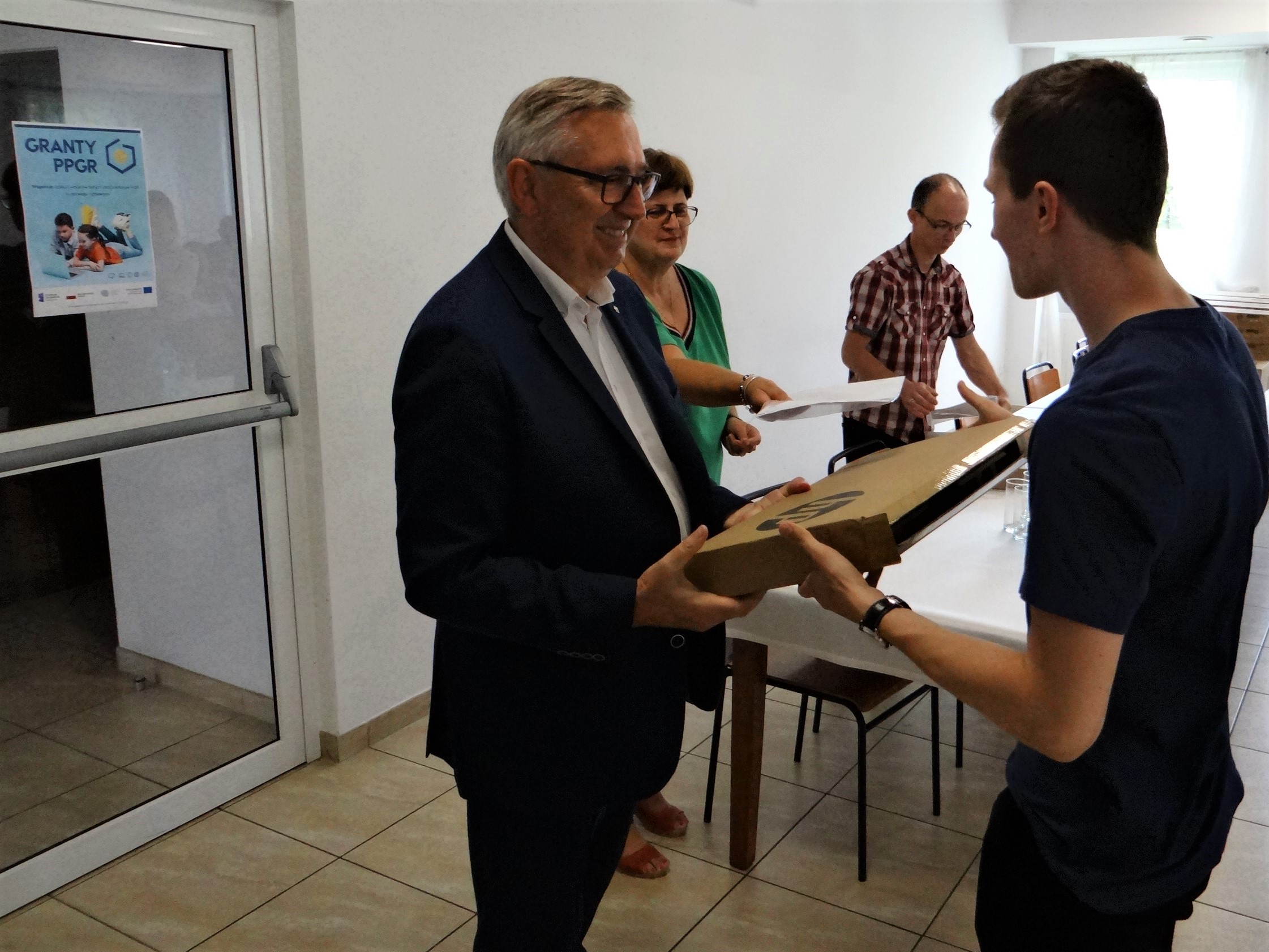 Wiceminister Stanisław Szwed przekazuje kolejnemu młodemu człowiekowi przenośny komputer. Wewnątrz budynku młodszy mężczyzna odbiera z rąk starszego opakowanie z laptopem w środku