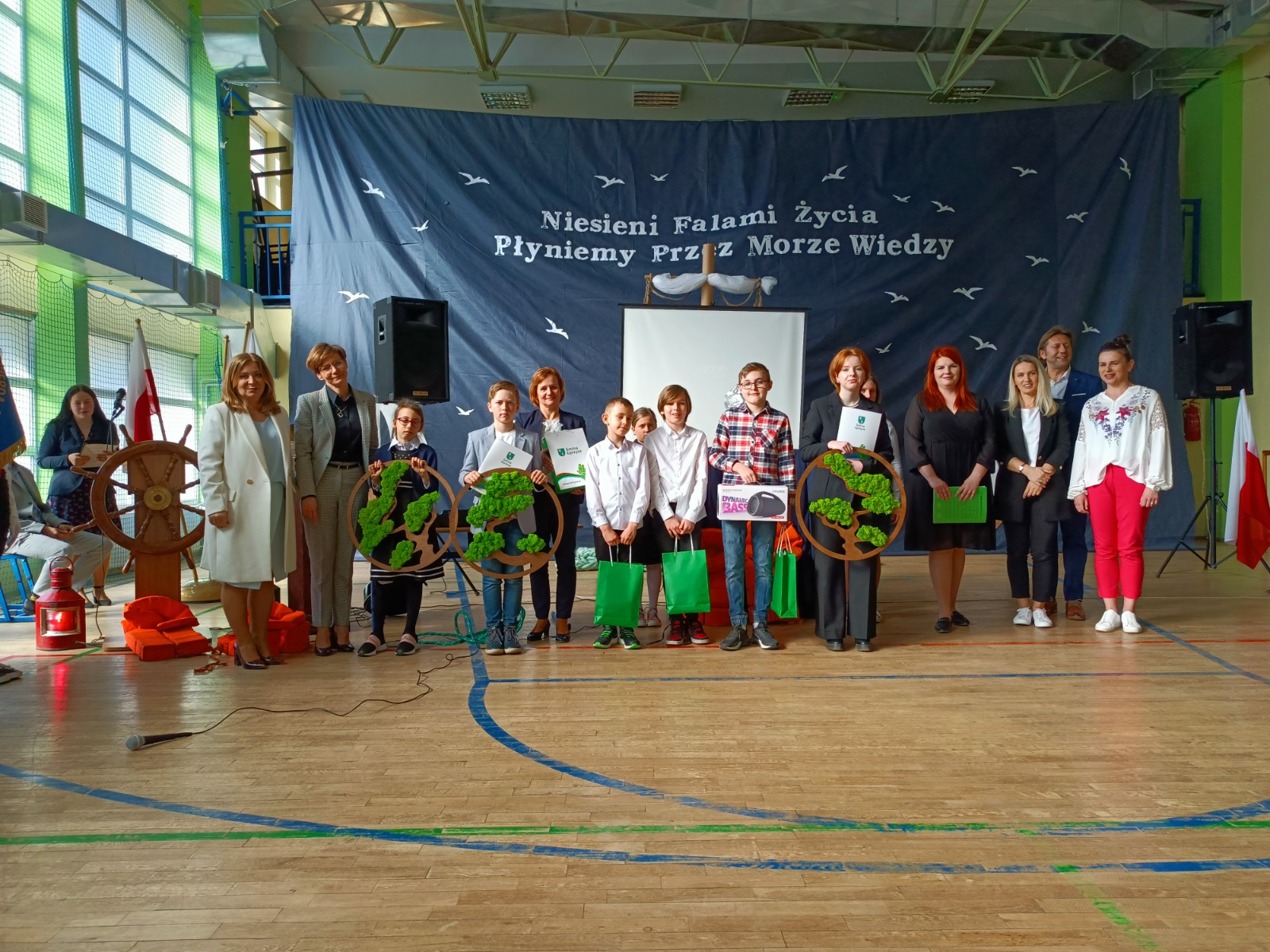 Zdjęcie przedstawiające dzieci i dorosłych na sali gimnastycznej na wręczeniu nagród w konkursie, w tle tekst: " Niesieni Falami Życia Płyniemy Przez Morze Wiedzy"