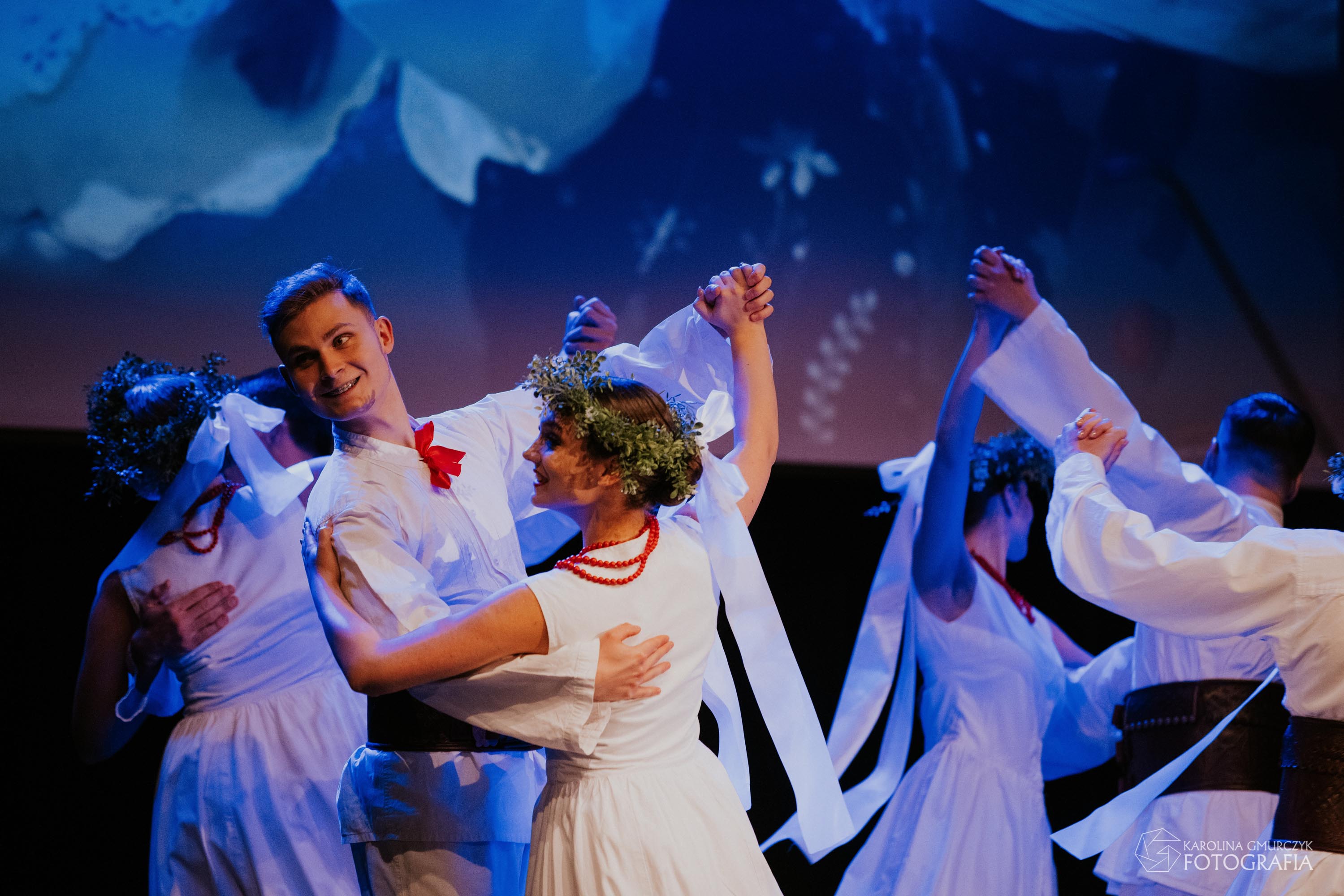 Na scenie Sokołowskiego Ośrodka Kultury widać wirujące w tańcu pary ubrane w tradycyjne regionalne stroje ludowe w kolorze białym. Tancerki ubrane w białe sukienki z czerwonymi akcentami i zielonym wiankiem na głowie. Tancerze ubrani w długie białe spodnie i białe kaftany z czerwoną wstążką pod szyją, przepasani przez biodra skórzanymi pasami.