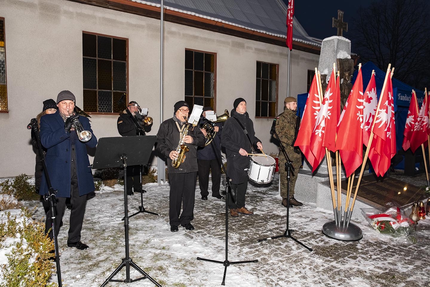 orkiestra stojąca na prawo od pomnika Powstańców Wielkopolskich, przed pomnikiem flagi Powstania Wielkopolskiego (biały orzeł na czerwonym tle)