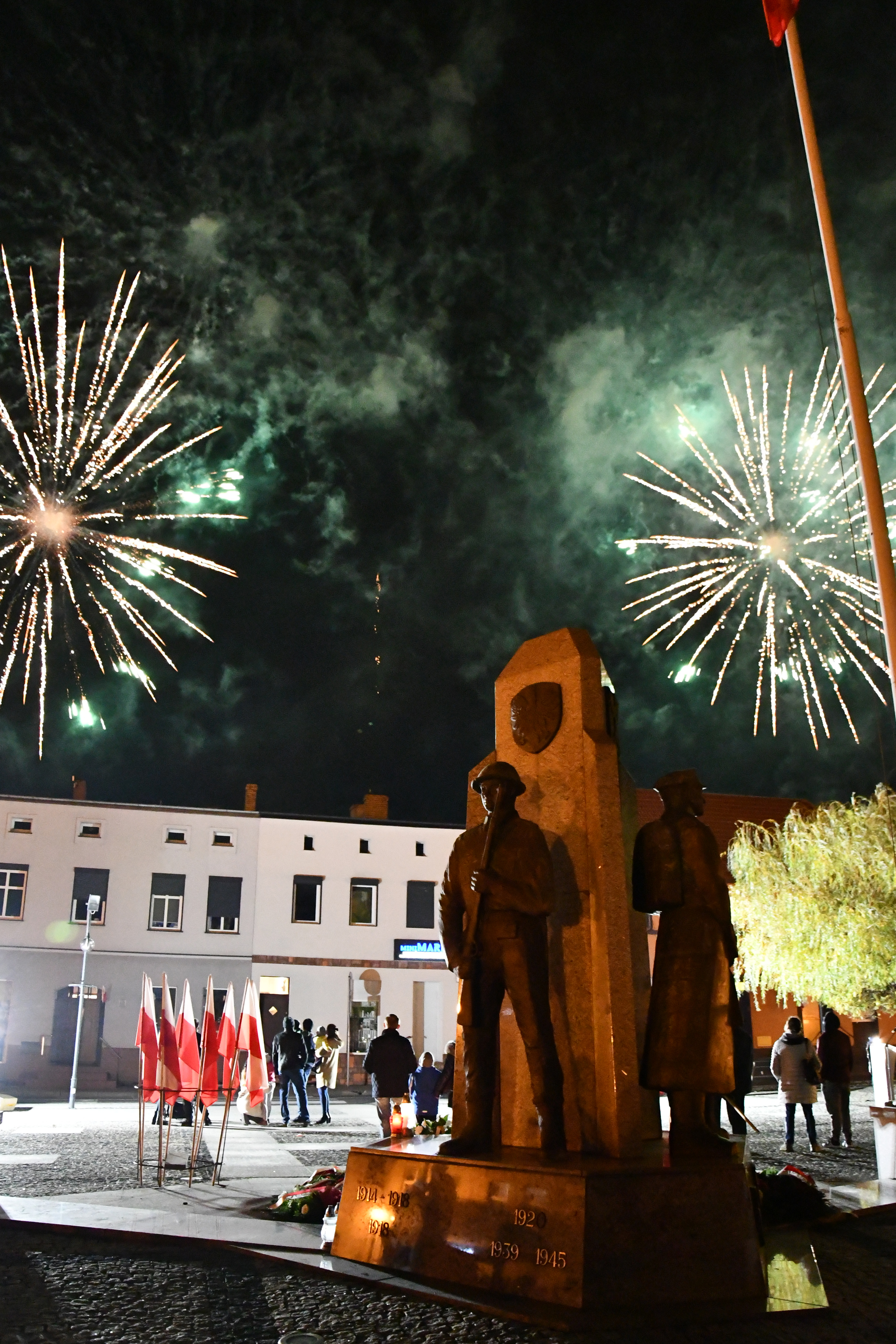 pokaz sztucznych ogni na niebie nad Rynkiem w Margoninie, na pierwszym planie pomnik Poległym za wolność