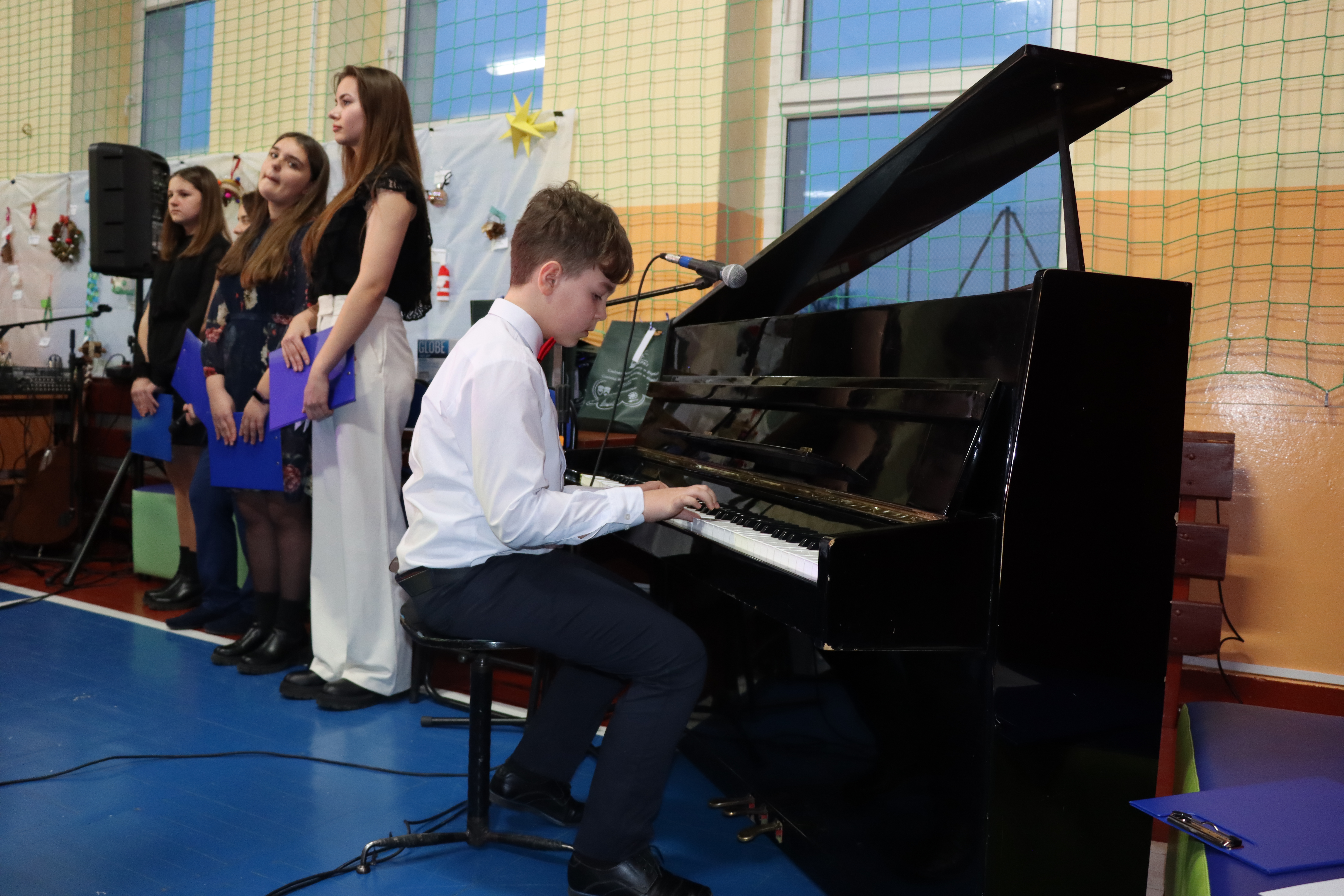 Chłopak ubrany na galowo (biała koszula, czarne spodnie wykonuje na pianinie pastorałkę, obok 4 osoby występujące w koncercie (recytujące)