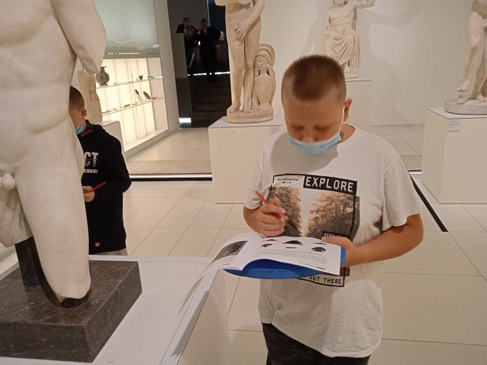 Chłopiec trzyma w rękach kartkę z napisami o obrazkami. Wokół niego stoją rzeźby ludzi