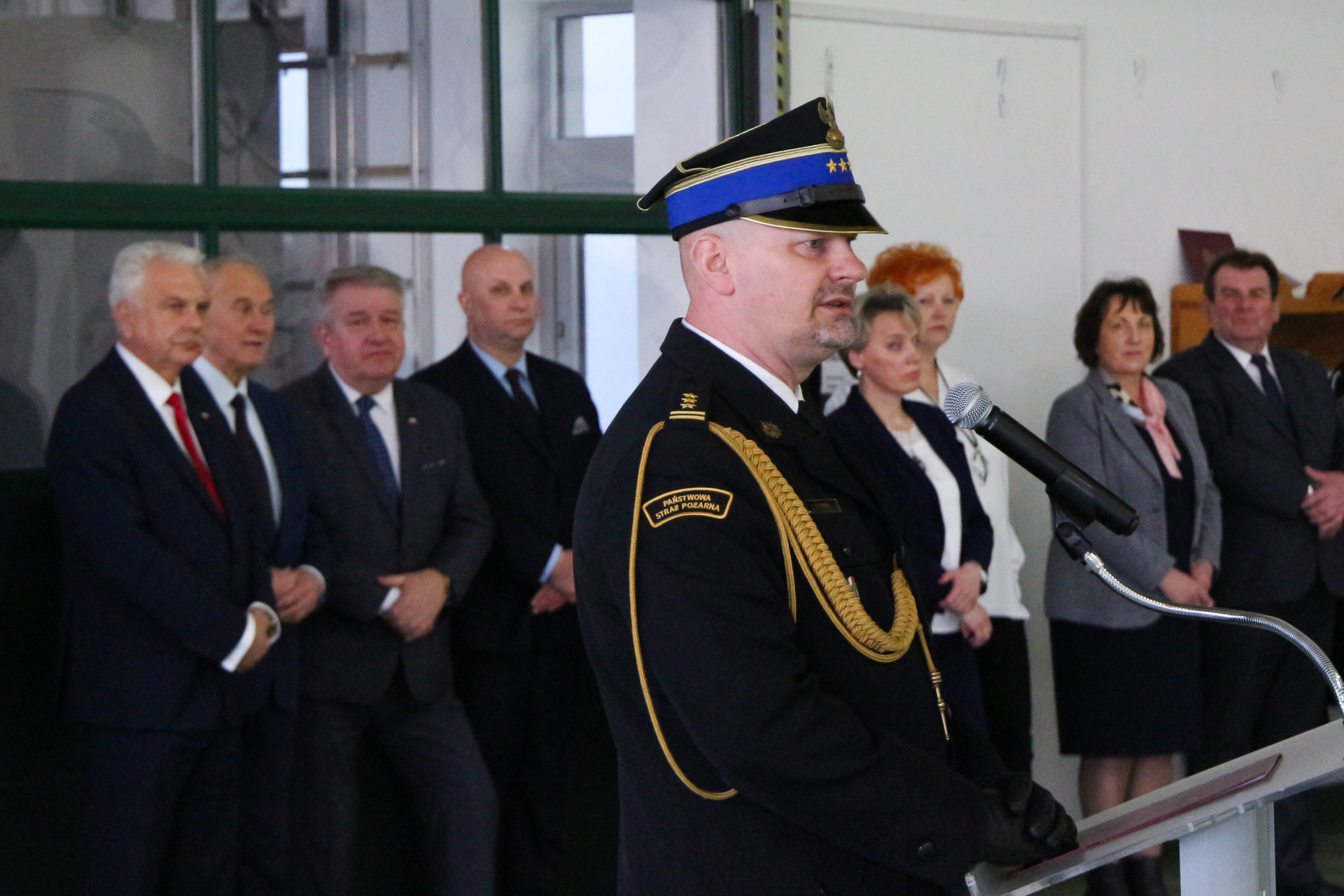 Na pierwszym planie przy metalowej mównicy z logo Państwowej Straży Pożarnej przemawia zastępca komendanta wojewódzkiego PSP w Warszawie, w tle stojący wzdłuż ściany hangaru goście.