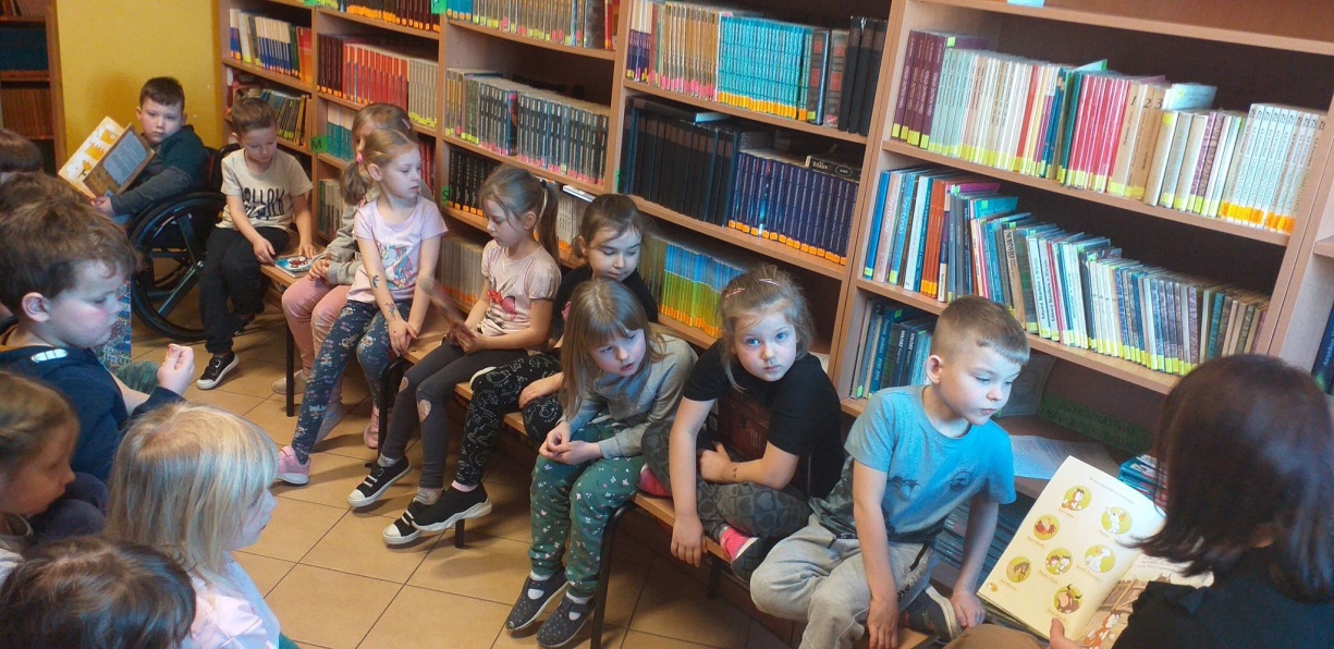 Dzieci siedzące na krzesłach. Za nimi stoją regały z książkami
