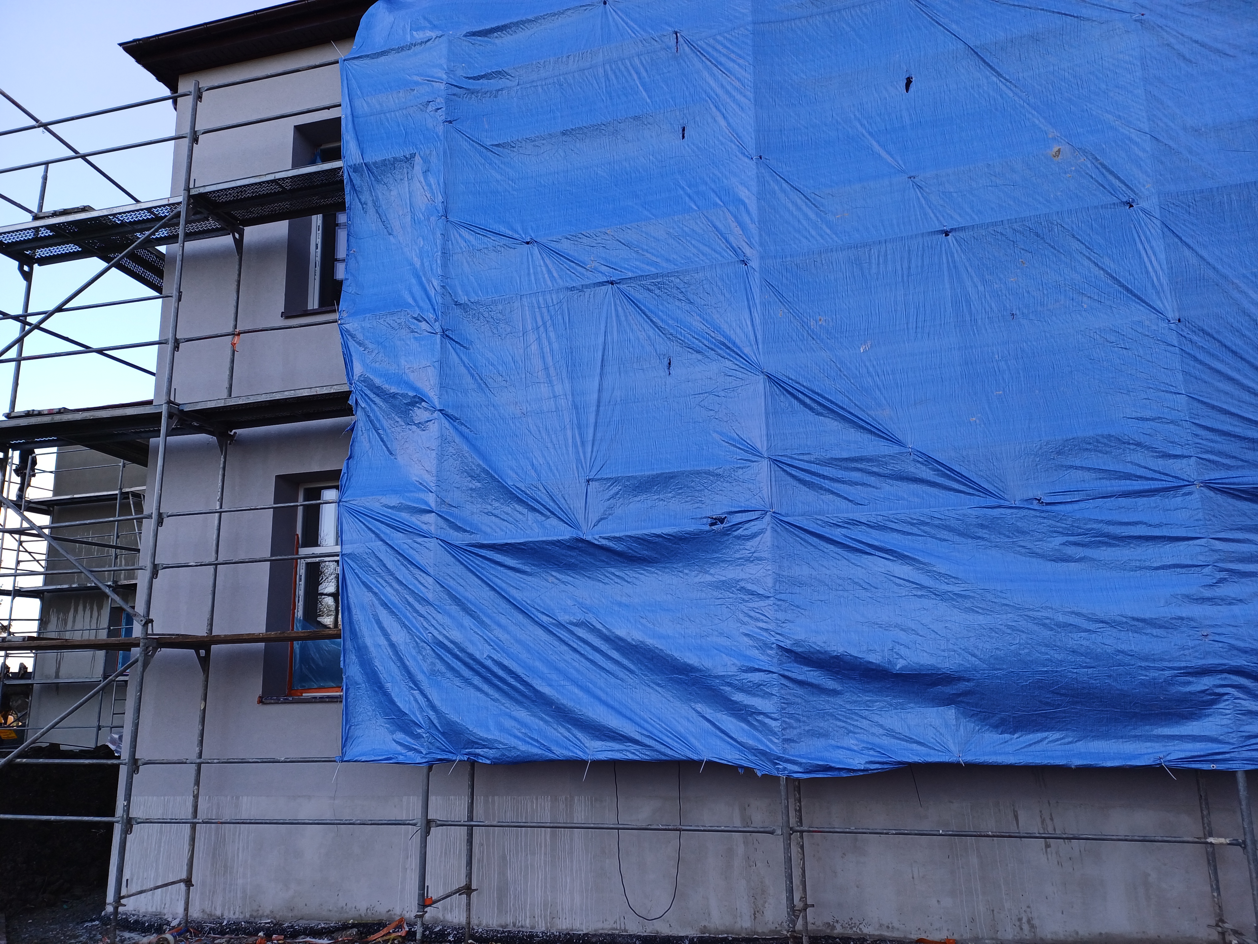 Zdjęcie przedstawia fragment budynku , obłożony rusztowaniem i częściowo zakryty niebieską plandeką.