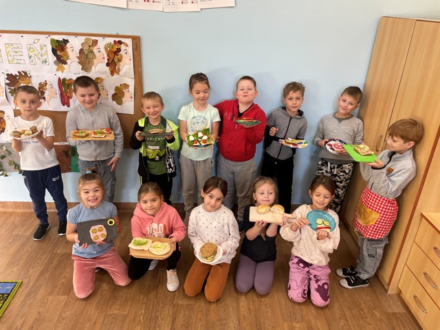 Fotografia przedstawia klasę pierwszą a,13- ro dzieci stoi w dwóch rzędach trzymając talerze ze zrobionymi kanapkami. Dzieci są uśmiechnięte i zadowolone ze swojej pracy.