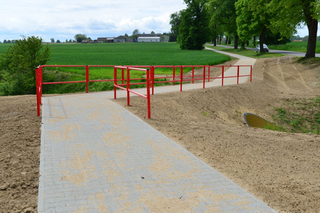 ścieżka rowerowa z kostki brukowej, nad przepustem z obu stron ścieżki czerwona metalowa barierka