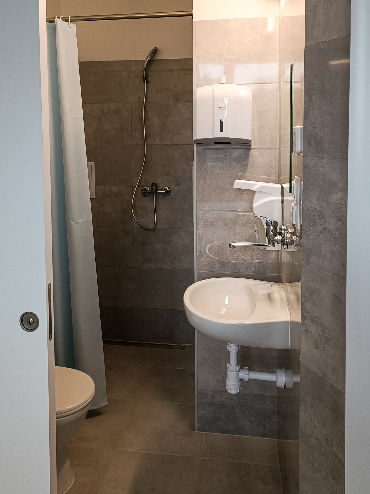 Na zdjęciu widoczne wyremontowane pomieszczenie łazienki, w której znajdują się prysznic, toaleta oraz umywalka, dozowniki środków dezynfekcyjnych i papieru