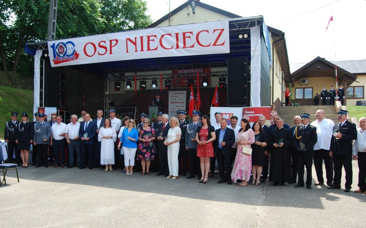 Widok na scenę. Zdjęcie grupowe ustawionych szeregach gości oraz Strażaków w mundurach galowych. Na zadaszeniu sceny biały baner z logiem OSP 100 lat w jedności i napisem OSP Nieciecz.