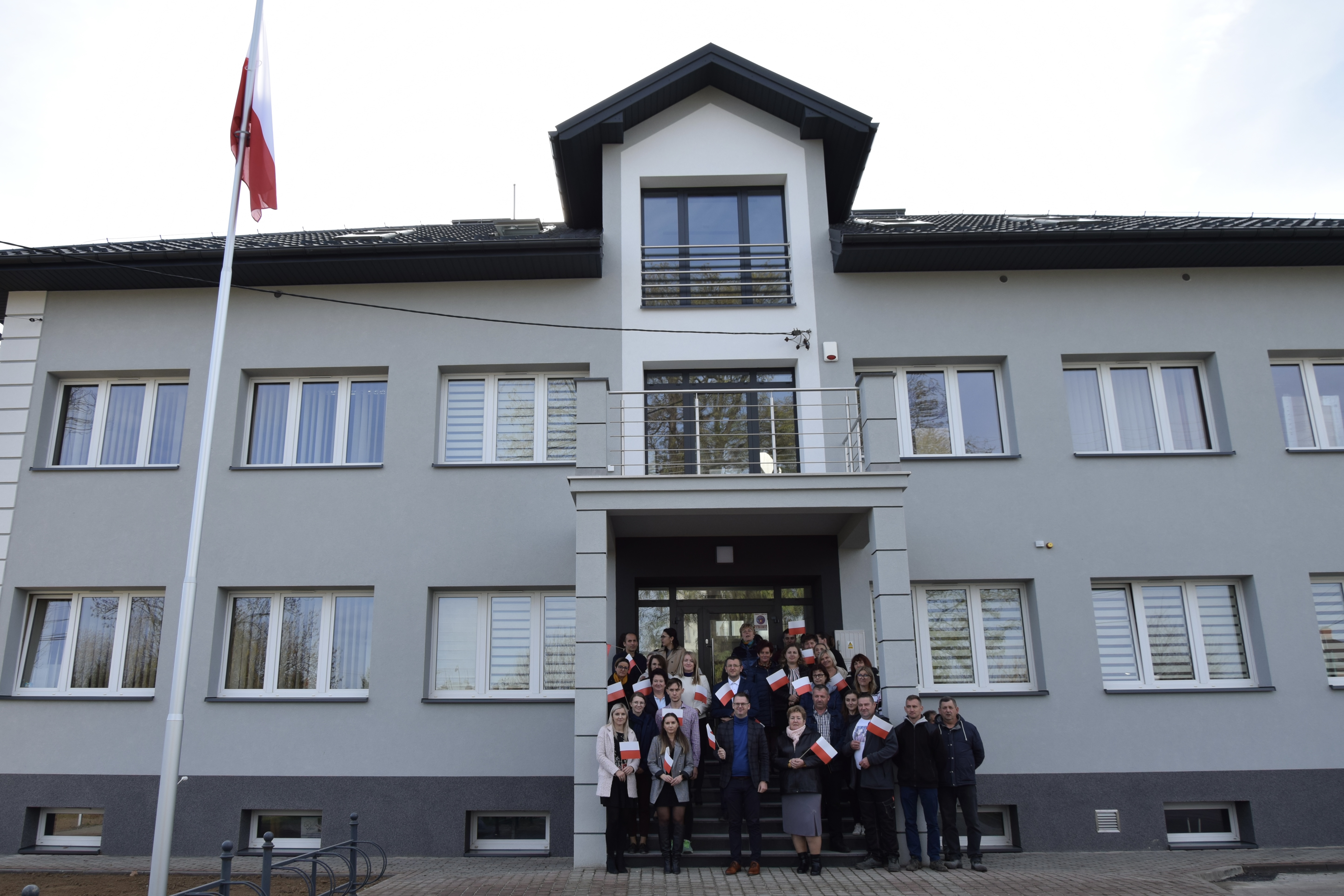 zdjęcie przedstawia pracowników urzędu na tle budynku Urzędu Gminy. Z lewej strony widnieje maszt z flagą Rzeczypospolitej Polskiej
