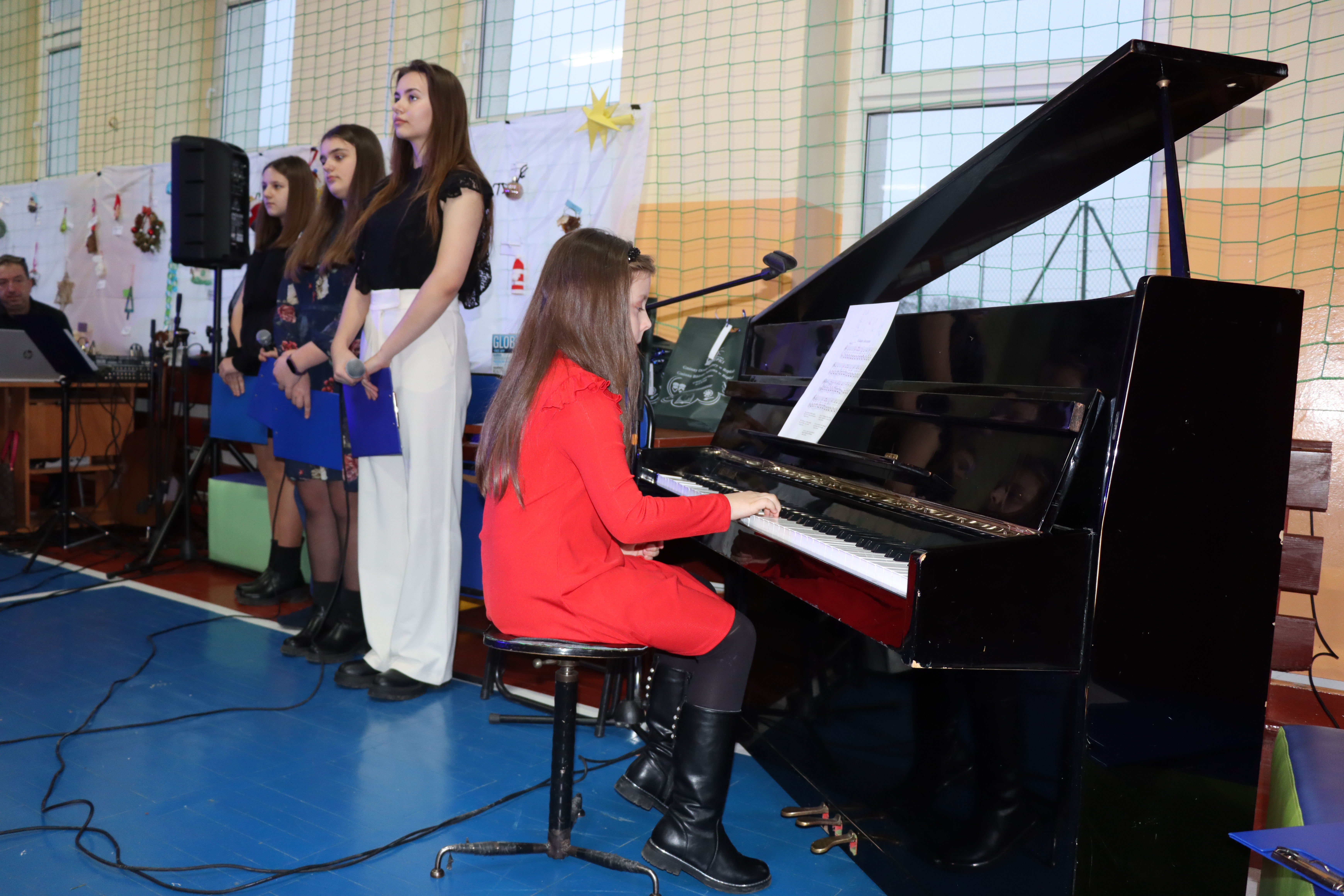 Dziewczynka w czerwonej sukience gra na pianinie jedną z kolęd, obok stoją cztery osoby, występujące w koncercie (recytujące)