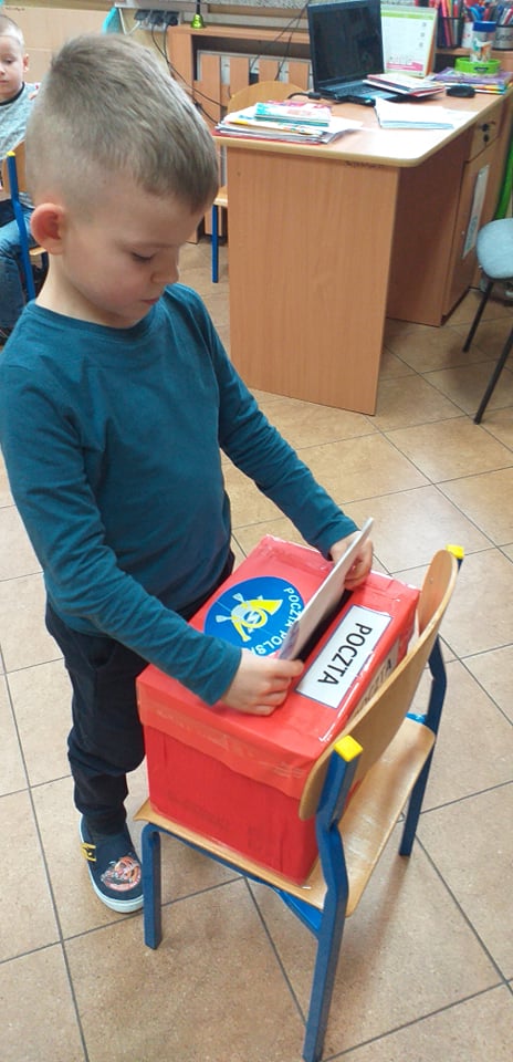 Chłopiec wkłada kartkę do czerwonej skrzynki z napisem Poczta