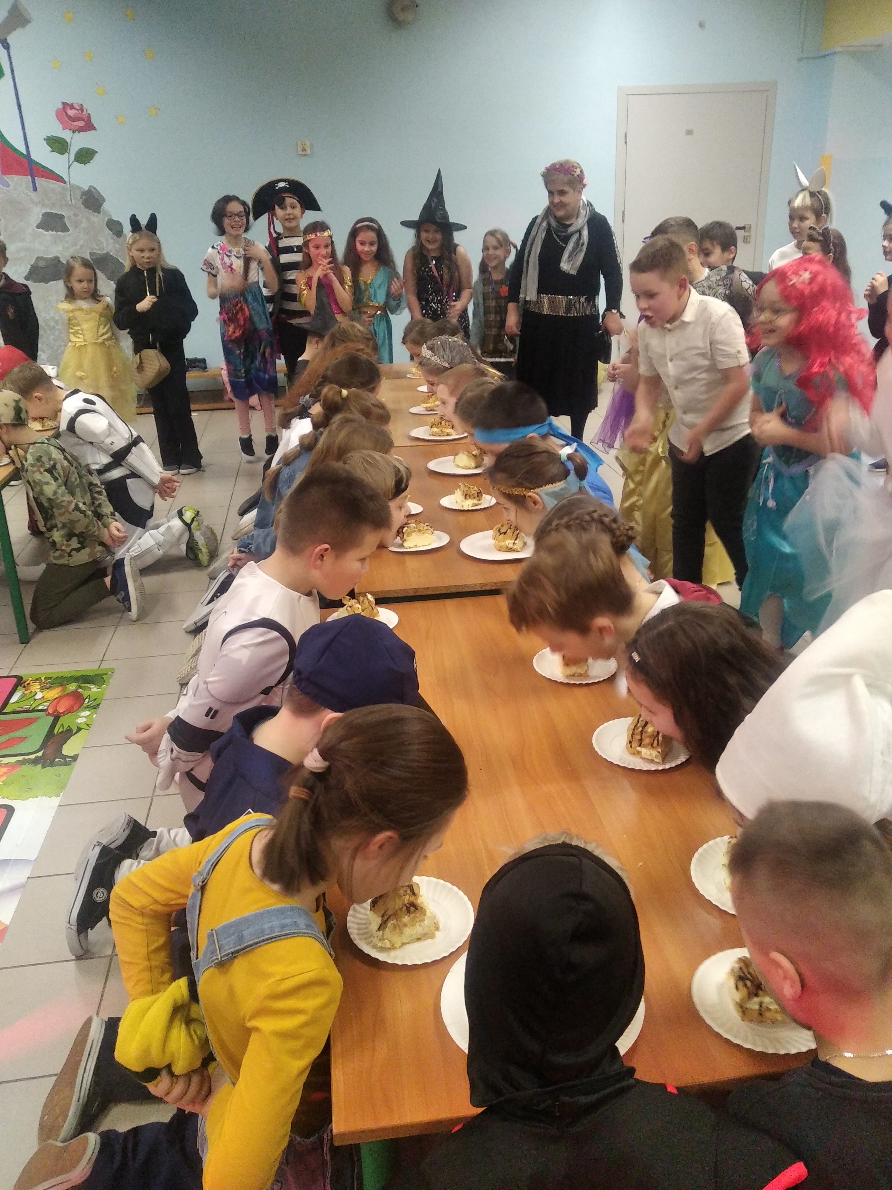 Dzieci pochylają głowę nad talerzyk z ciastkiem z kremem. Ręce mają schowane za plecami. Próbują zjeść pyszne ciastko. 
