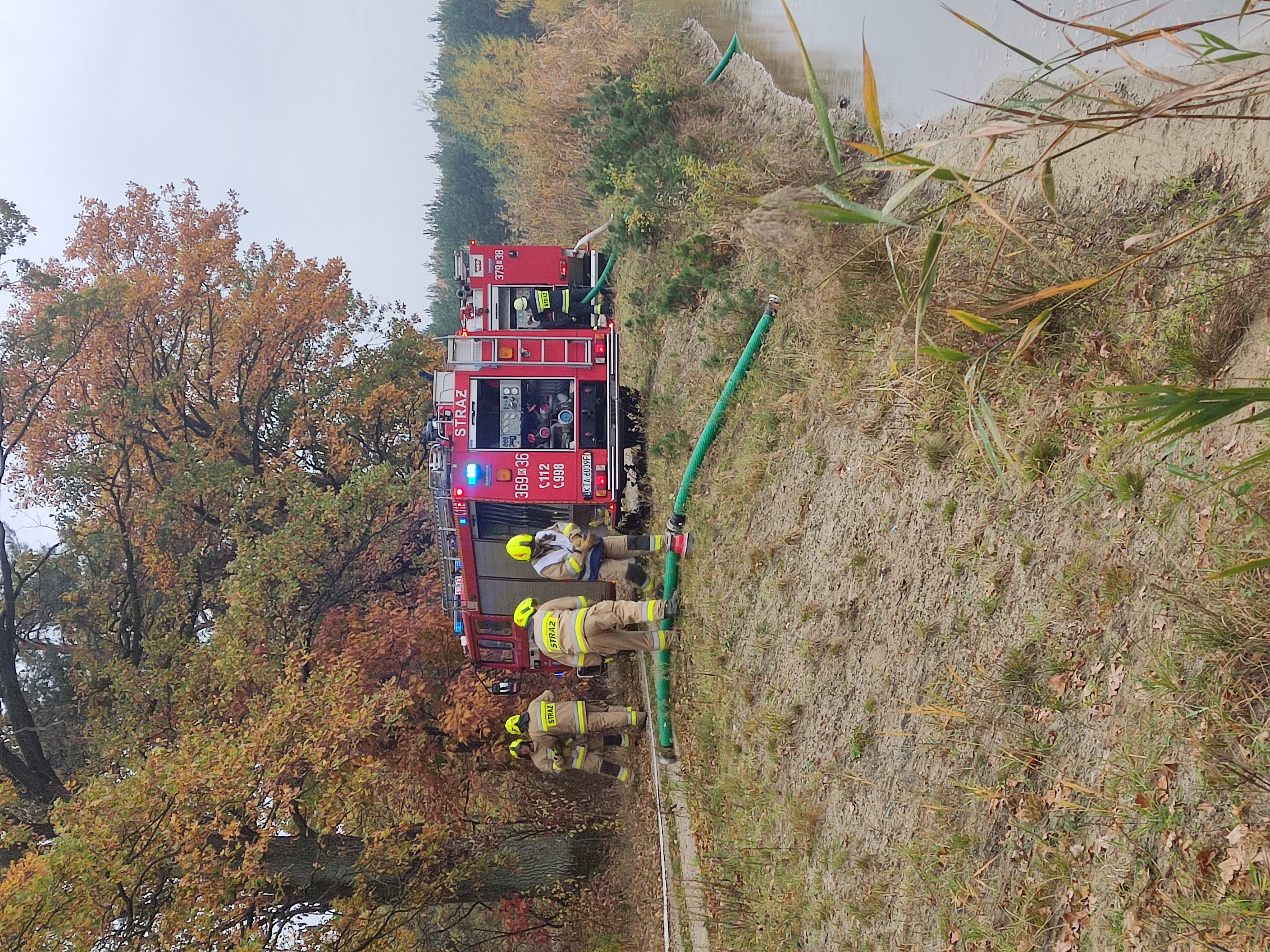 zdjęcie przedstawia dwa duże wozy strażackie oraz strażaków wraz ze sprzętem gaśniczym w trakcie ćwiczeń w lesie.