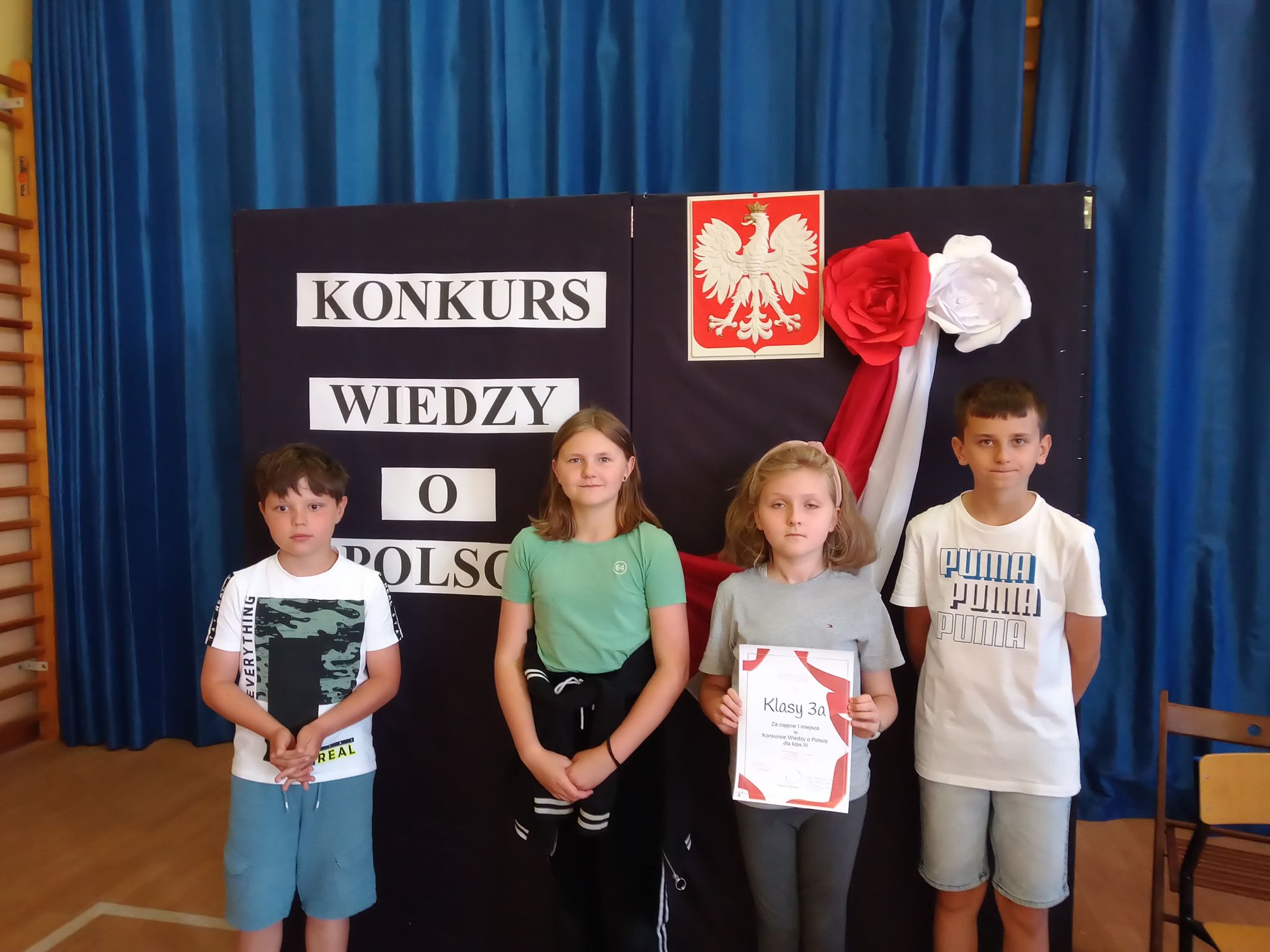 Czworo dzieci. Po środku dziewczynka trzyma w rękach dyplom. Za nimi godło Polski i napis Konkurs wiedzy o Polsce