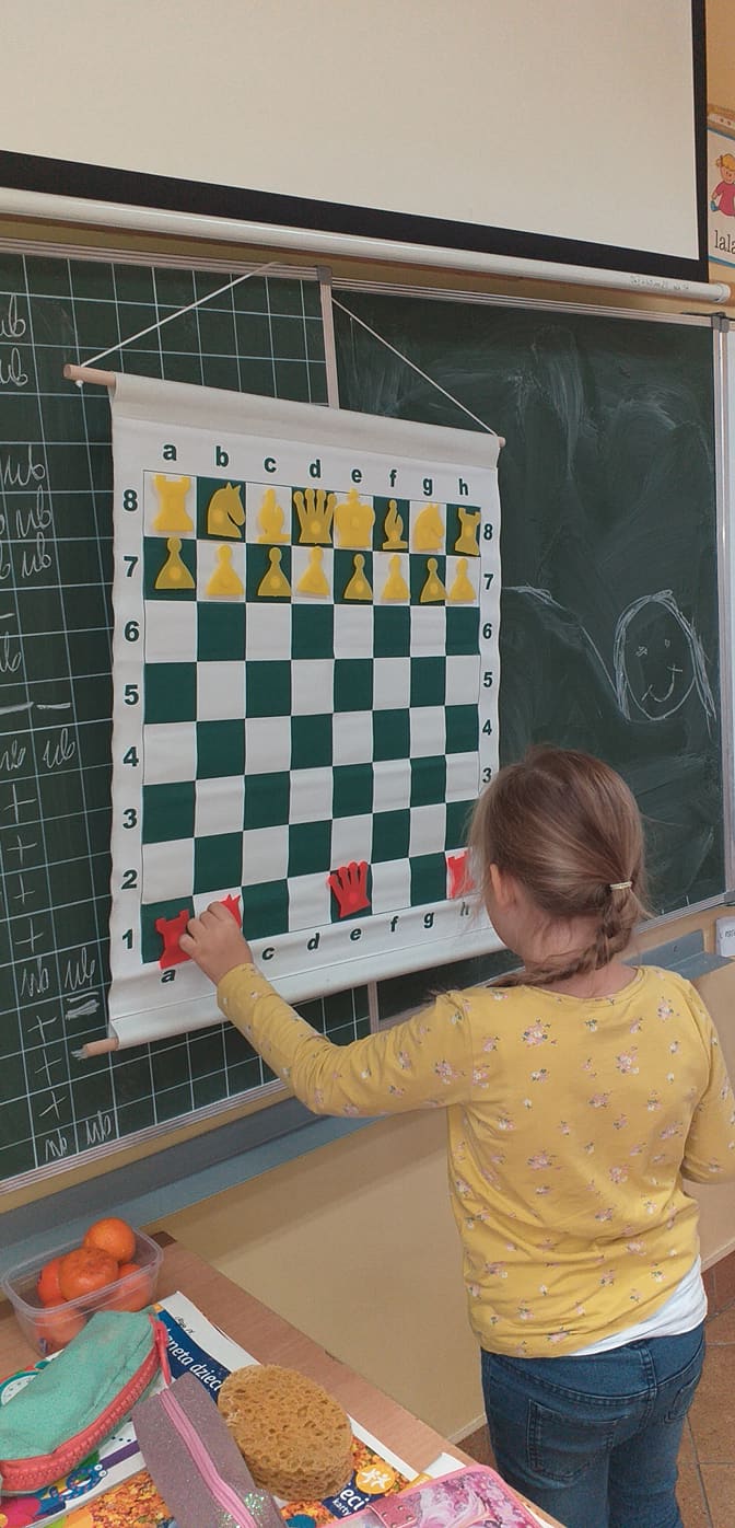 Dziewczynka stoi przy wiszącej szachownicy. Lewą ręką przesuwa pion