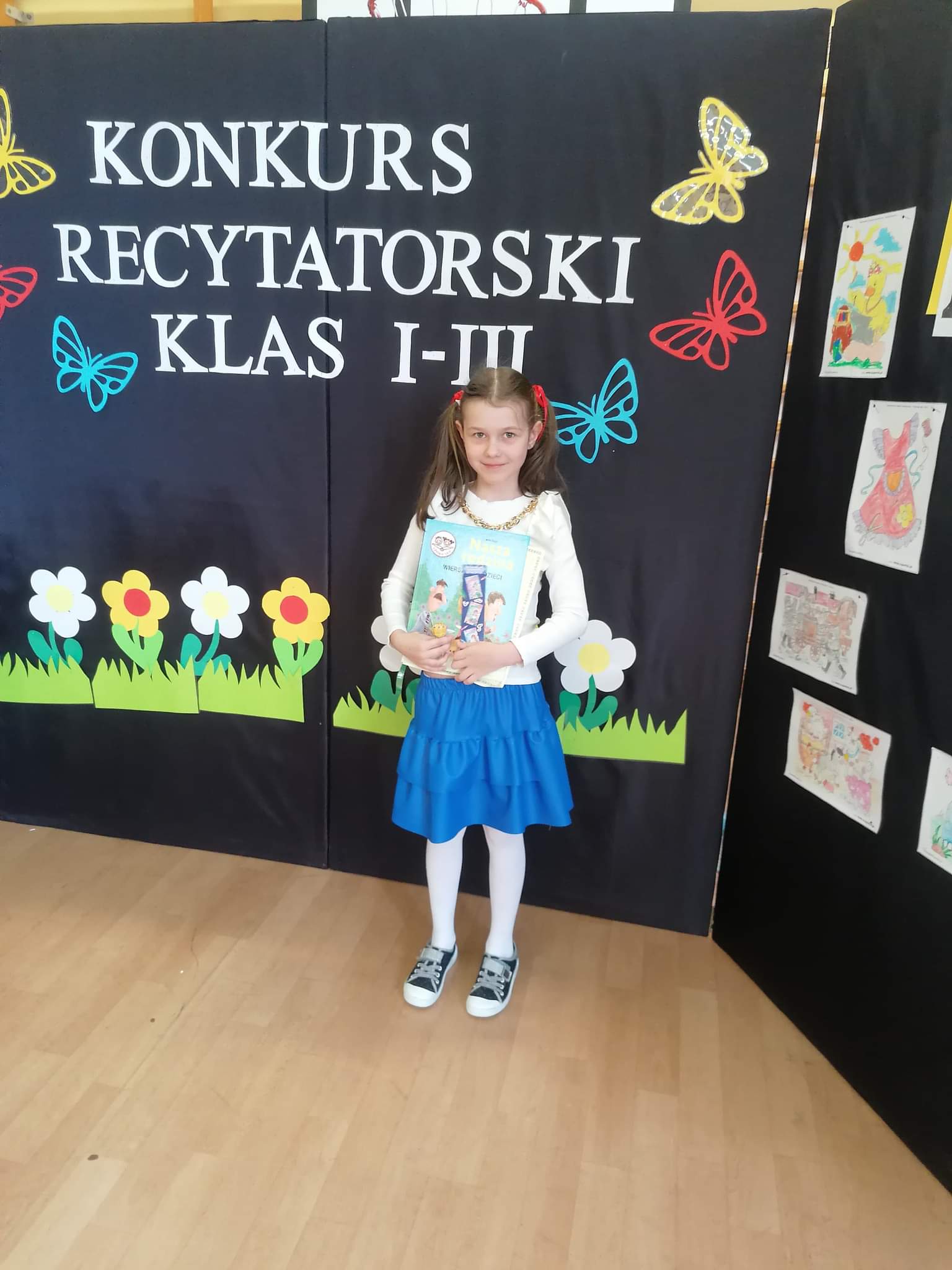 Dziewczynka trzymająca książkę w rękach stoi a za nią tablica z tekstem Konkurs recytatorski klas I-III