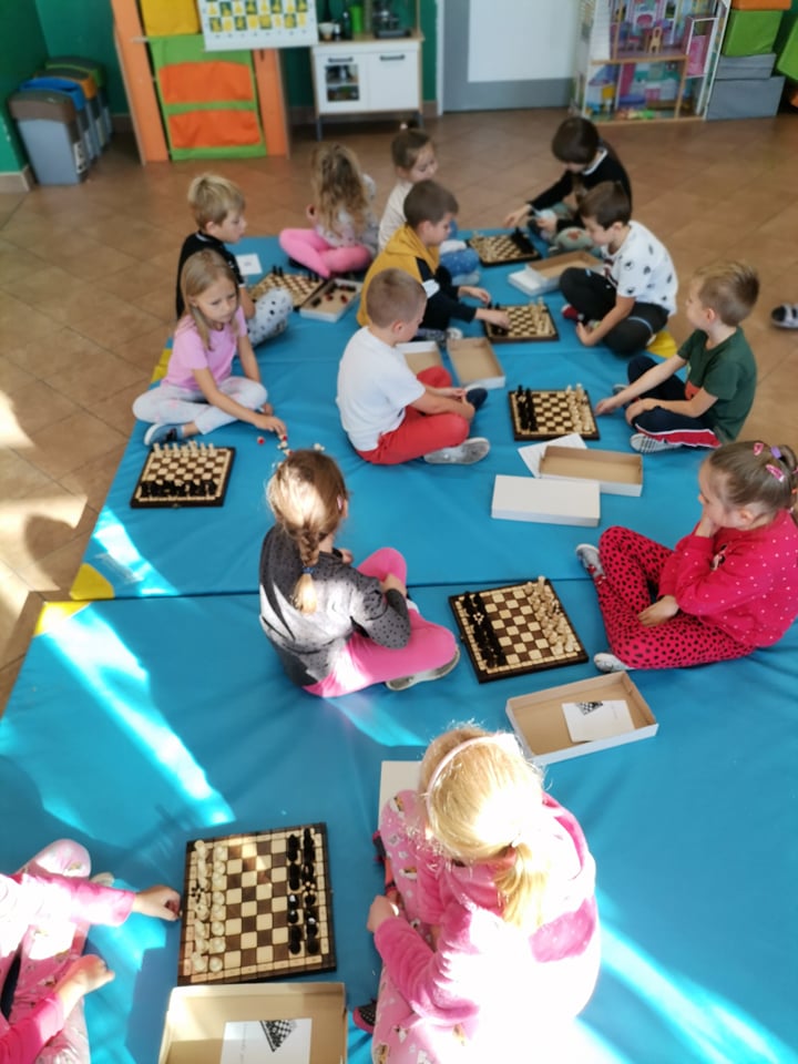 Dzieci siedzą w parach na niebieskim materacu. Pomiędzy nimi leżą szachownice