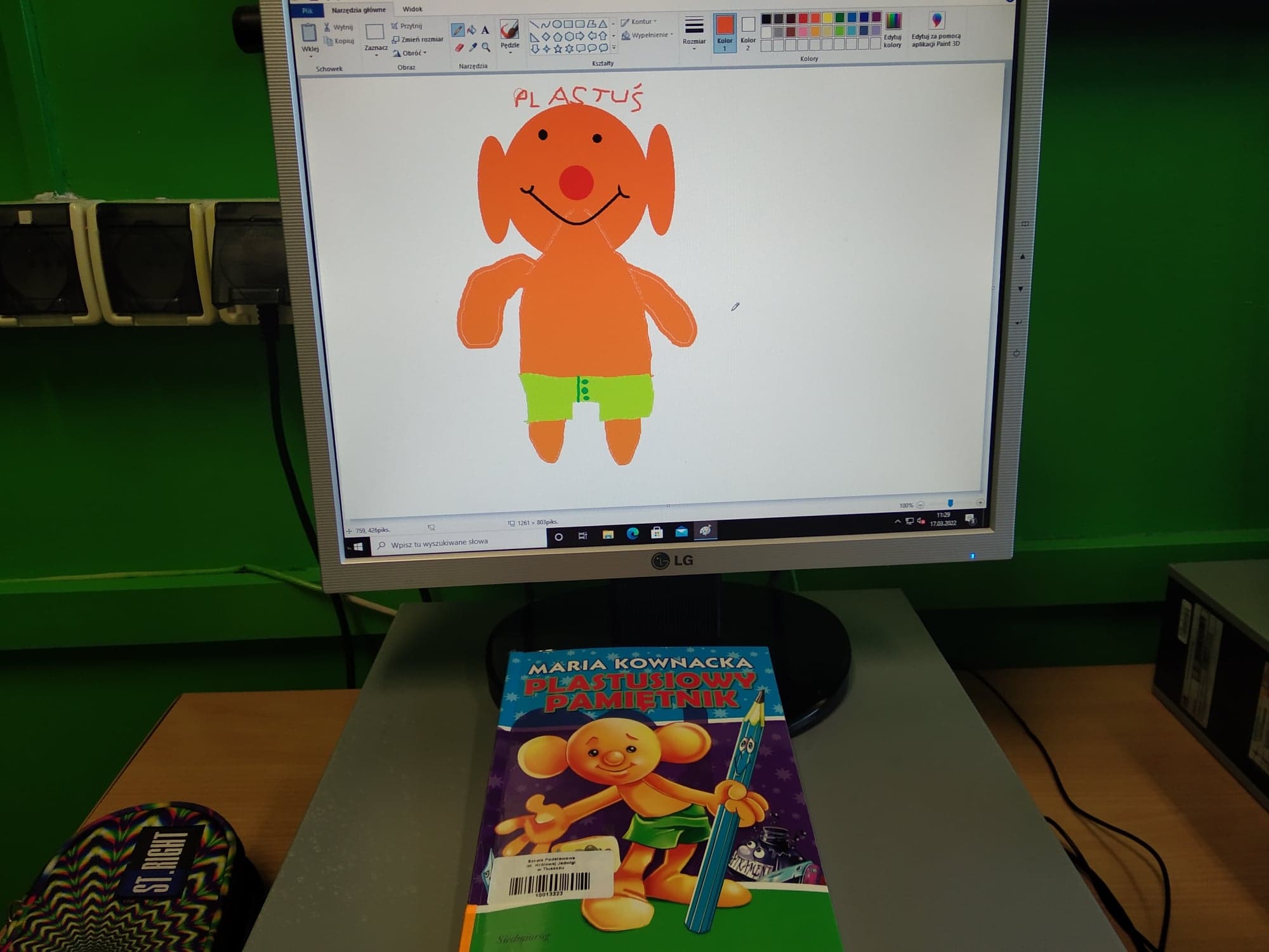 Pomarańczowa postać z dużymi uszami, z żółtymi szortami na ekranie komputera. Poniżej książka z okładką z pomarańczową postacią