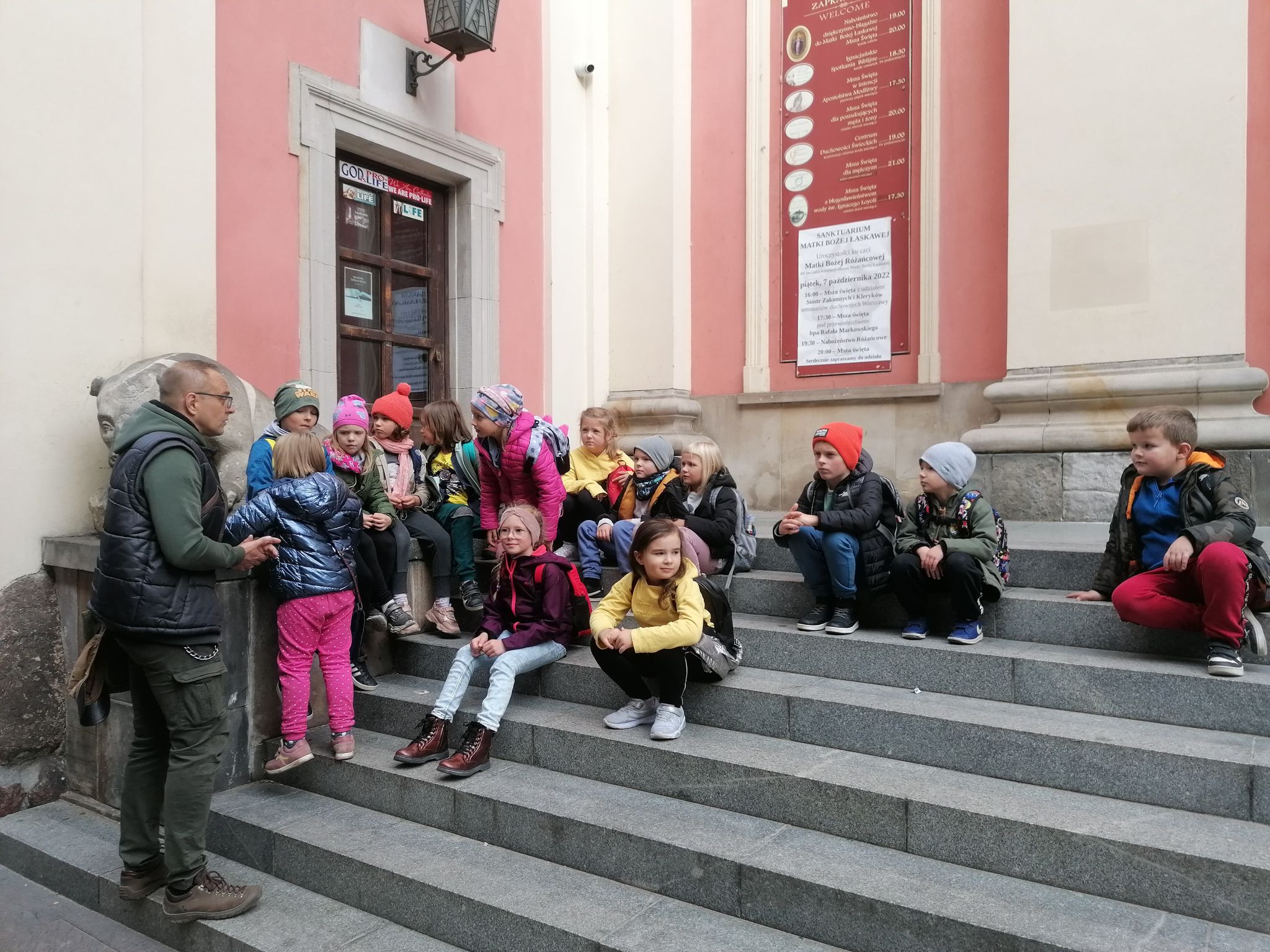 Dzieci siedzą na schodach przy budynku. Przed nimi stoi mężczyzna