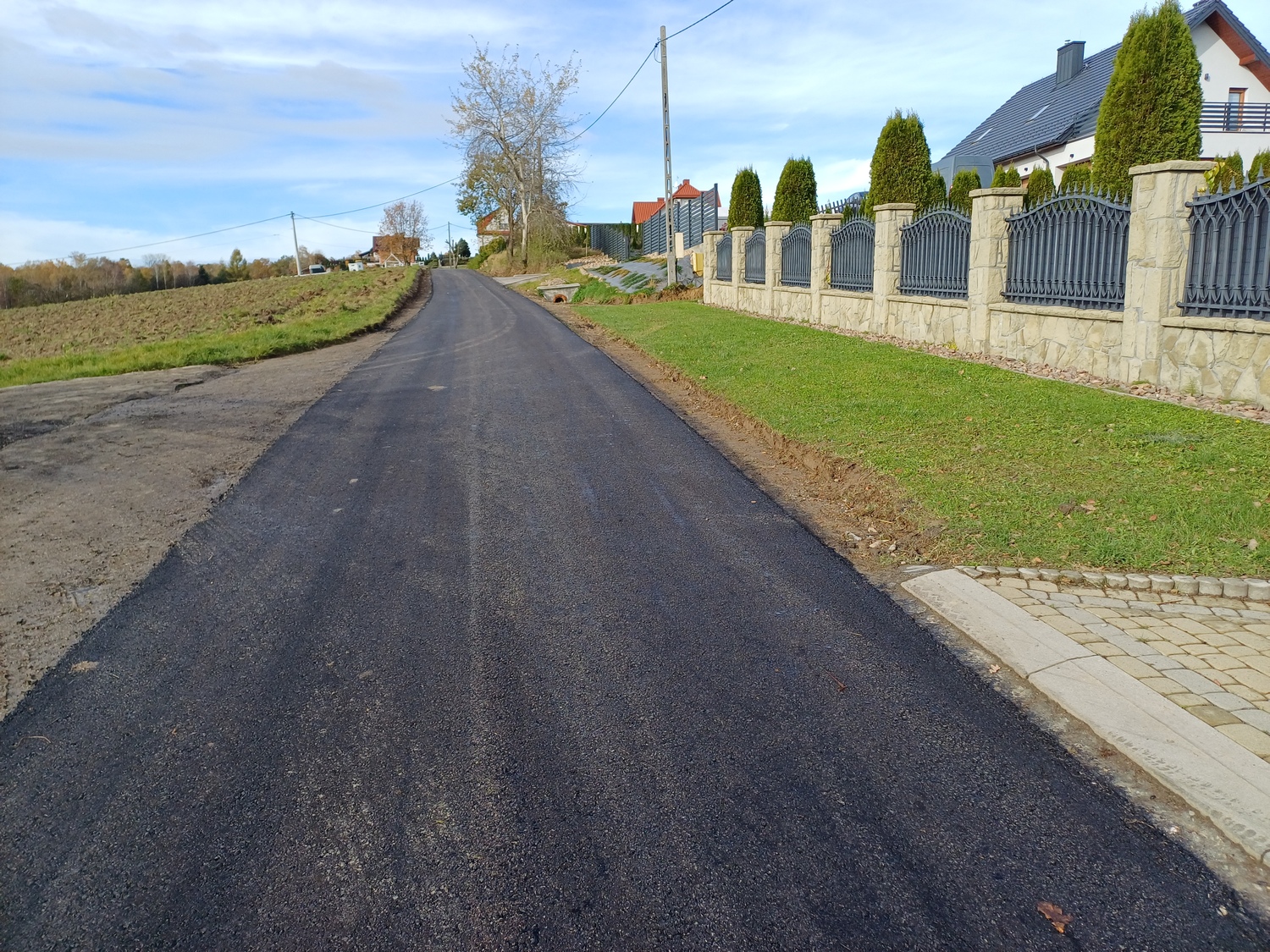 Fotografia przedstawia świeżo wyasfaltowaną drogę lokalną przebiegającą przez teren zabudowany. Po prawej stronie znajduje się ogrodzenie posesji z eleganckim murem i zadbanym trawnikiem, a po lewej pola uprawne. Droga jest równa, z wyraźnie uformowanymi poboczami i nowym wjazdem do posesji.