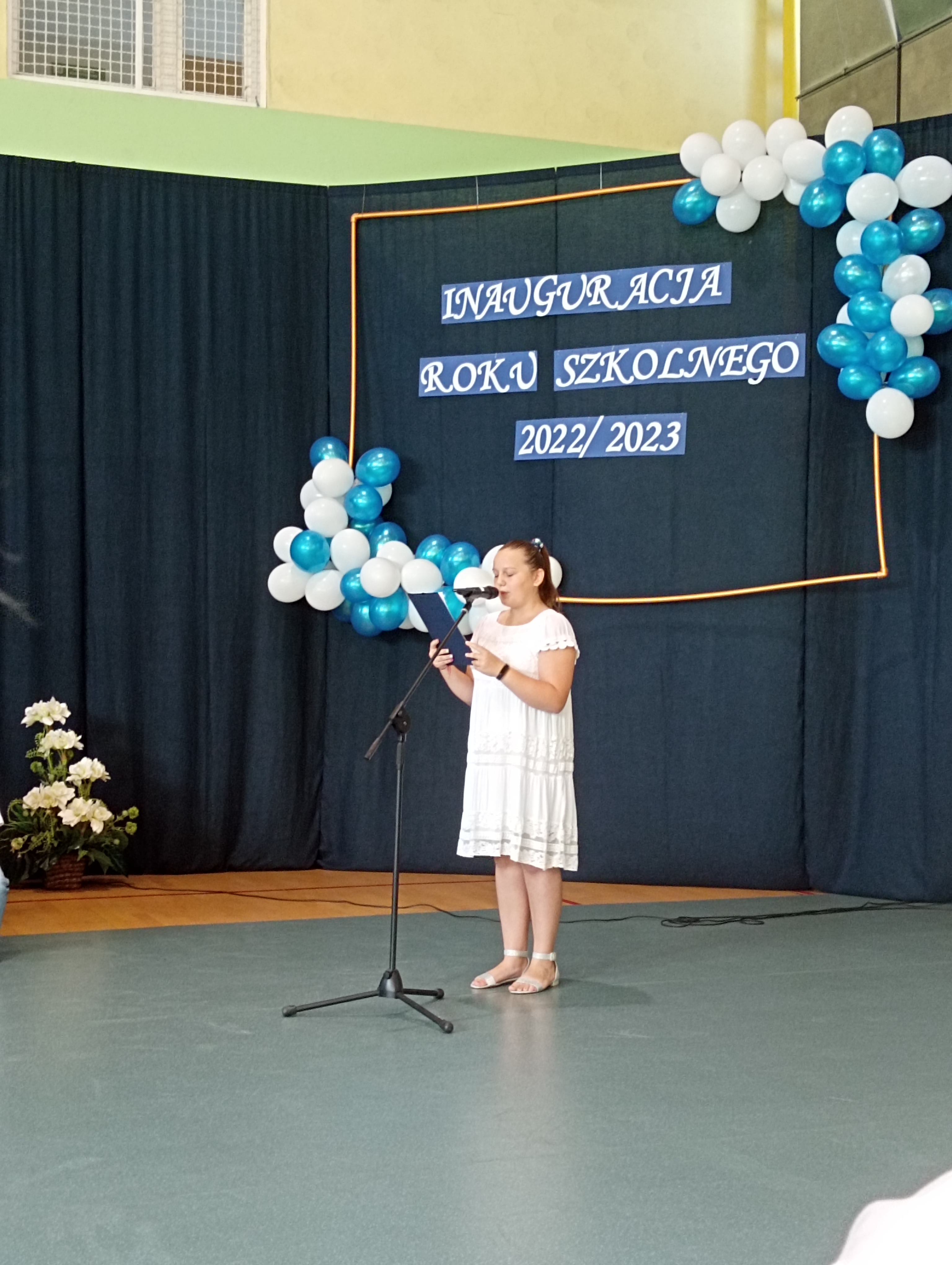 Dziewczynka w białej sukience trzyma w ręku niebieską teczkę i mówi do mikrofonu