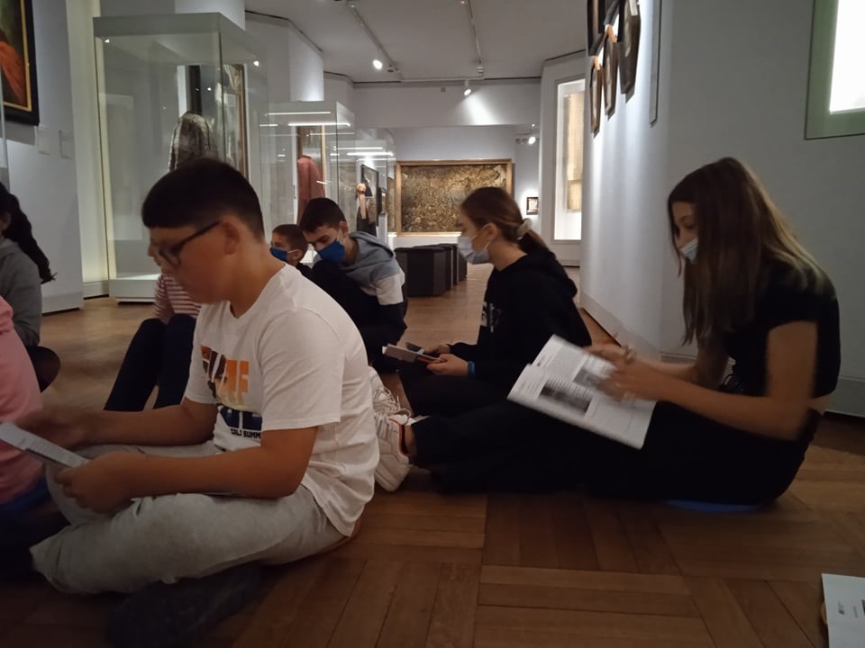 Młodzież siedzi na podłodze i czyta broszury. Za nimi stoją szklane gabloty