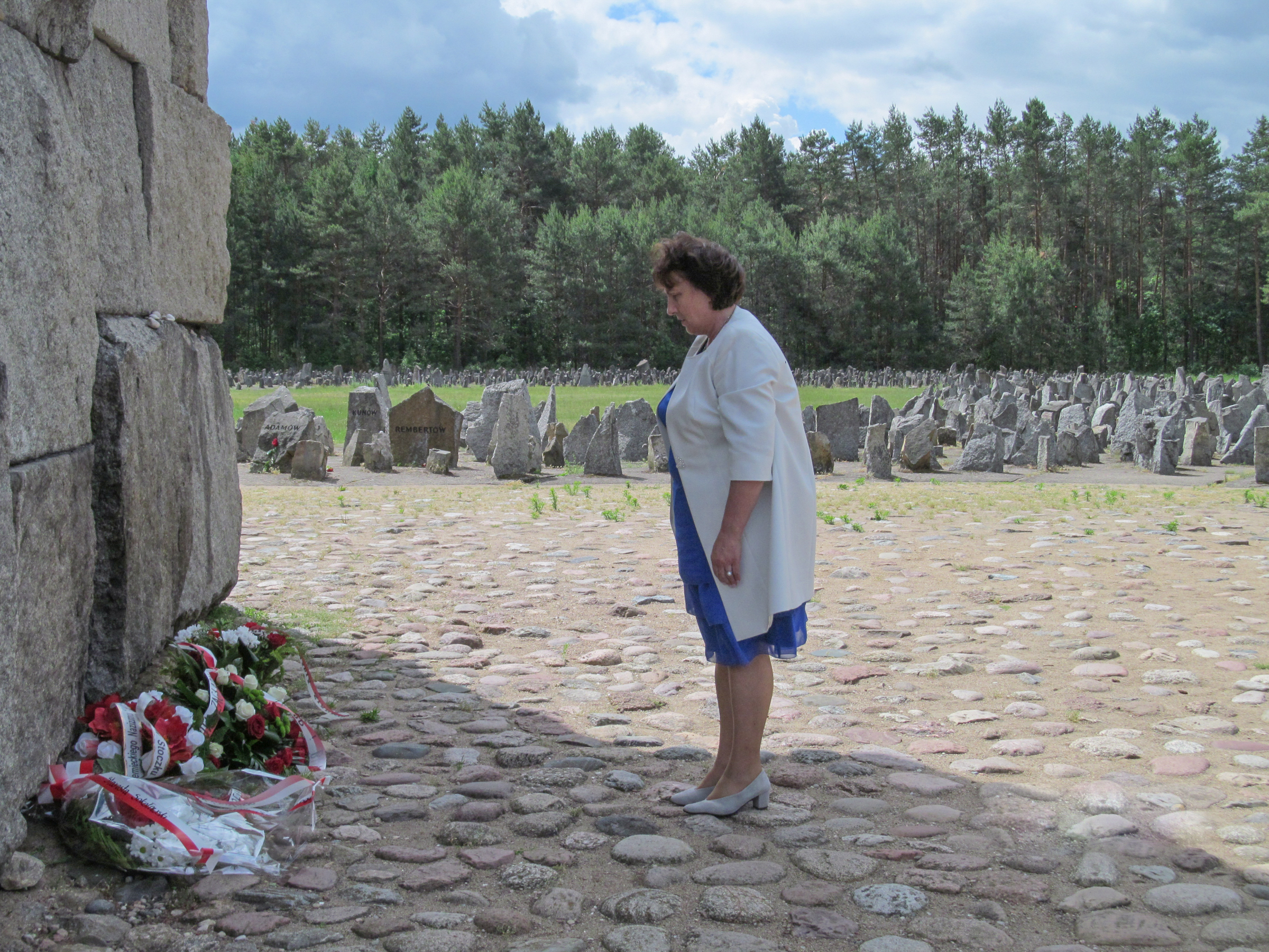 Fotografia przedstawia widok z boku na Starostę Elżbietę Sadowską, która stoi przed głównym monumentem Ofiar Obozu Zagłady w Treblince. W tle setki granitowych głazów upamiętniających Ofiary. W oddali widać ścianę lasu.