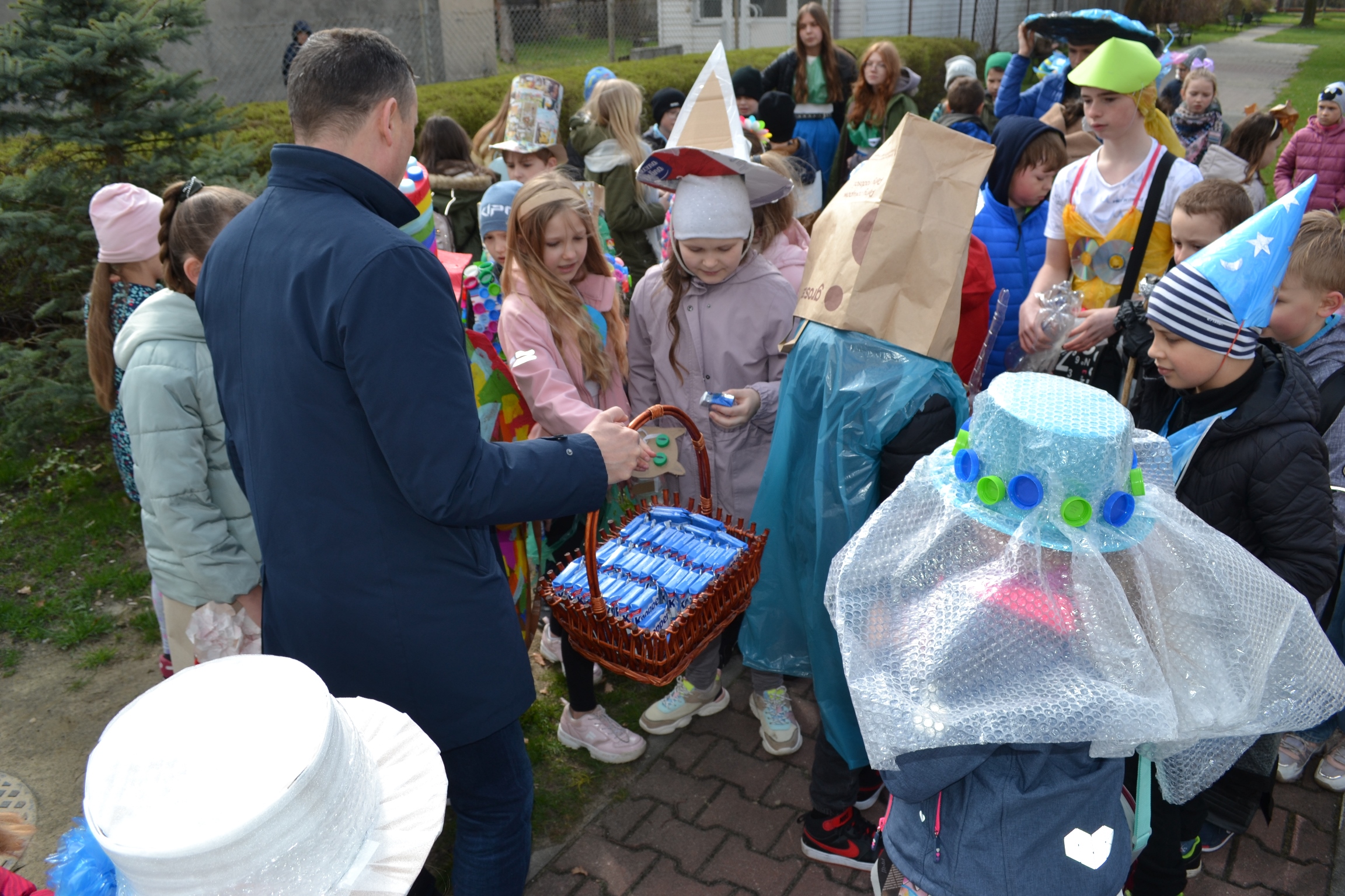 Mężczyzna trzyma koszyk z niebieskimi paczkami słodyczy. Przed nim stoją dzieci w kolorowych strojach
