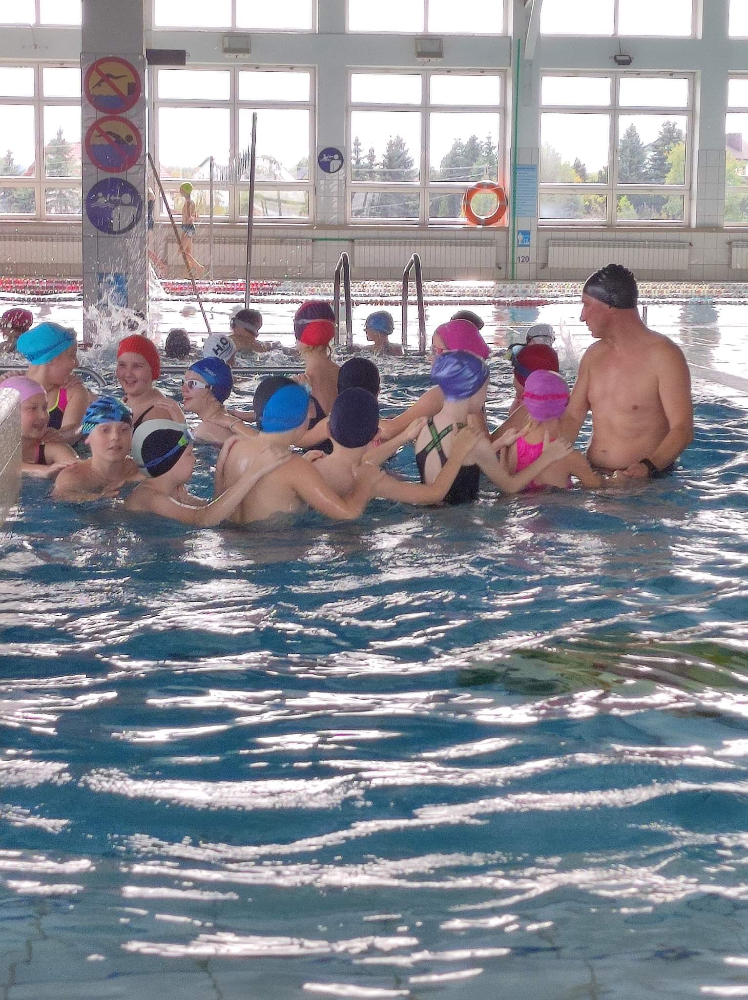 zdjęcie przedstawia grupkę dzieci w wieku szkolnym wraz z instruktorem pływania