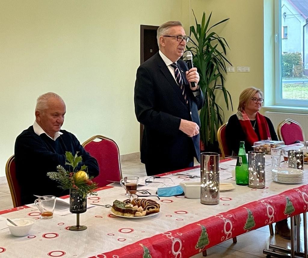 Minister Stanisław Szwed zwraca się do członków Klubu Seniora. Za stołem prezydialnym znajdują się trzy osoby, minister mówi do mikrofonu na stojąco.