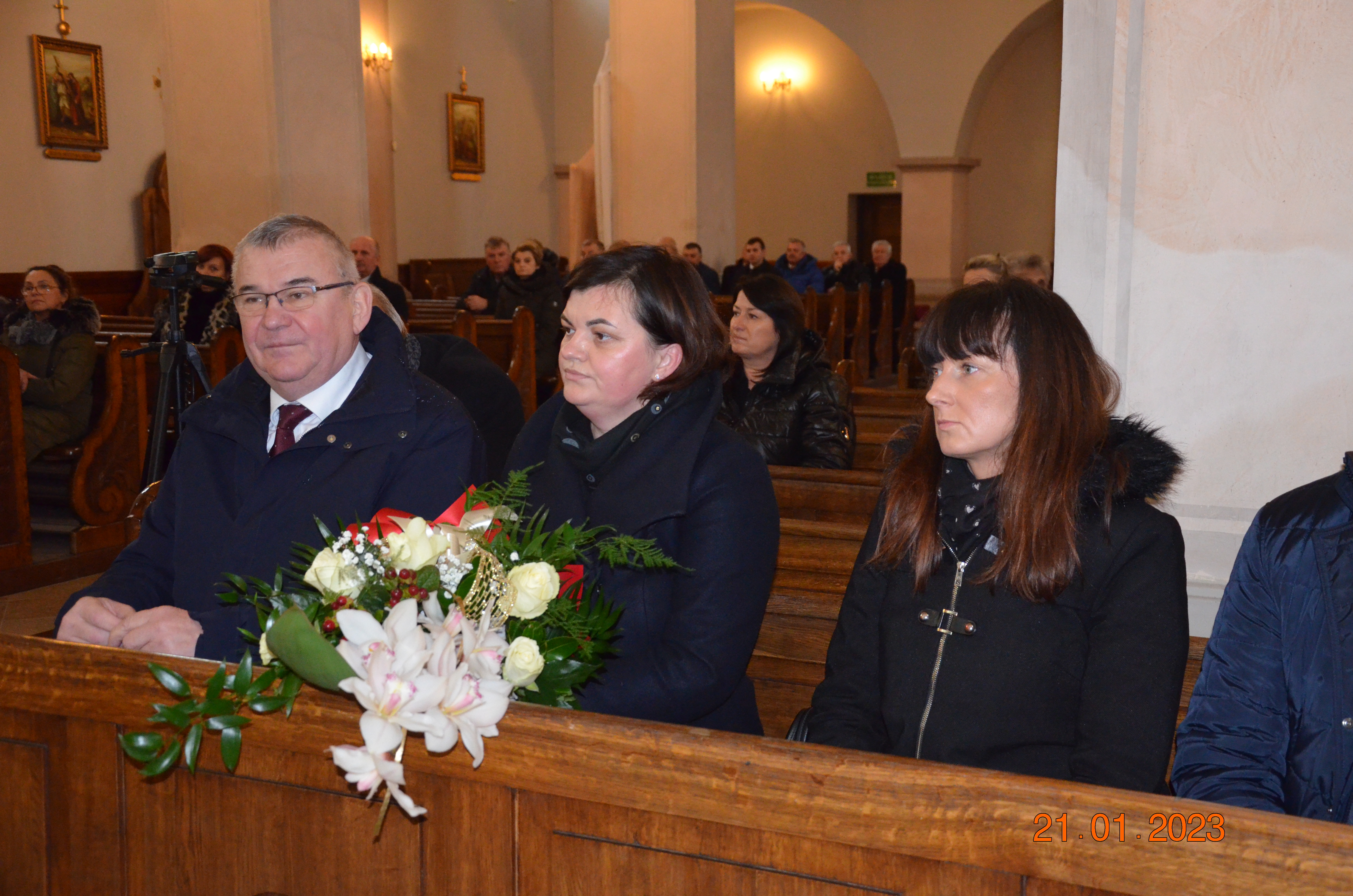 Mężczyzna oraz dwie kobiety siedzący w kościelnej ławie, kobieta trzyma w rękach bukiet kwiatów.