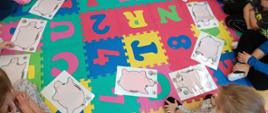 Dzieci z oddziału przedszkolnego siedzą na podłodze, na kolorowych matach rozłożone są rysunki skarbonki-świnki, na które dzieci kładą pieniążki