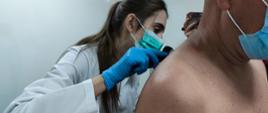 Festyn Profilaktyczno Zdrowotny w ogrodach przasnyskiego szpitala. Zdjęcie przedstawia wnętrze Dermatobusa oraz lekarkę w białym kitlu, niebieskich rękawiczkach i seledynowej maseczce dokonującej badania skóry pleców mężczyzny siedzącego przed nią.