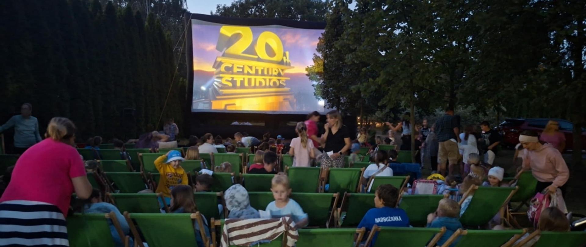 Grupa ludzi siedzi na leżakach na polance pod drzewami. Przed nimi duży ekran prokcyjny