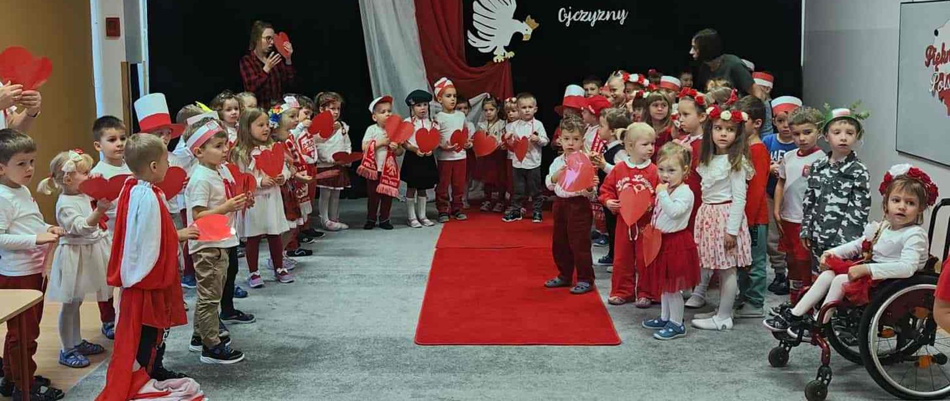 Dzieci w biało-czerwonych strojach trzymają czerwone serca i śpiewają piosenkę.