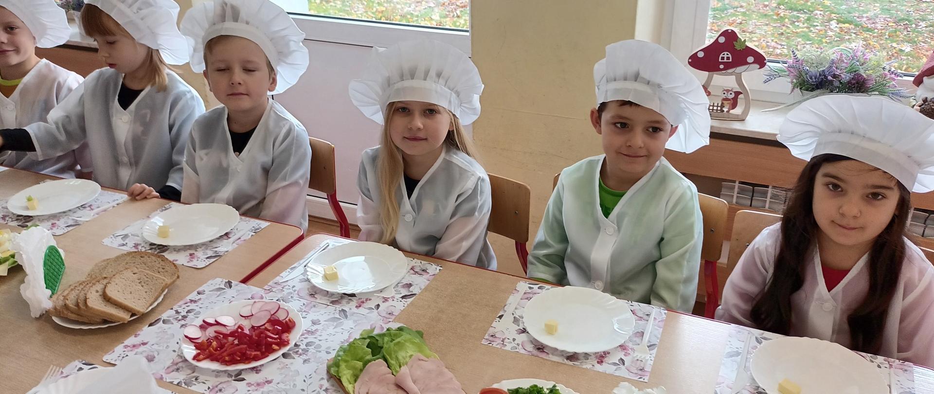 Dzieci w strojach kucharzy siedzą przy stole z posiłkiem 