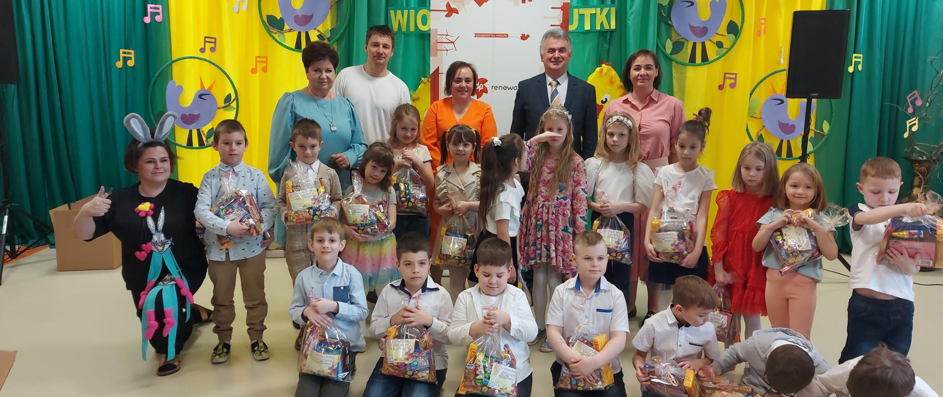 grupa dzieci częściowo klęczących, częściowo stojących, za nimi 6 dorosłych stojących na tle zielono-żółtych dekoracji