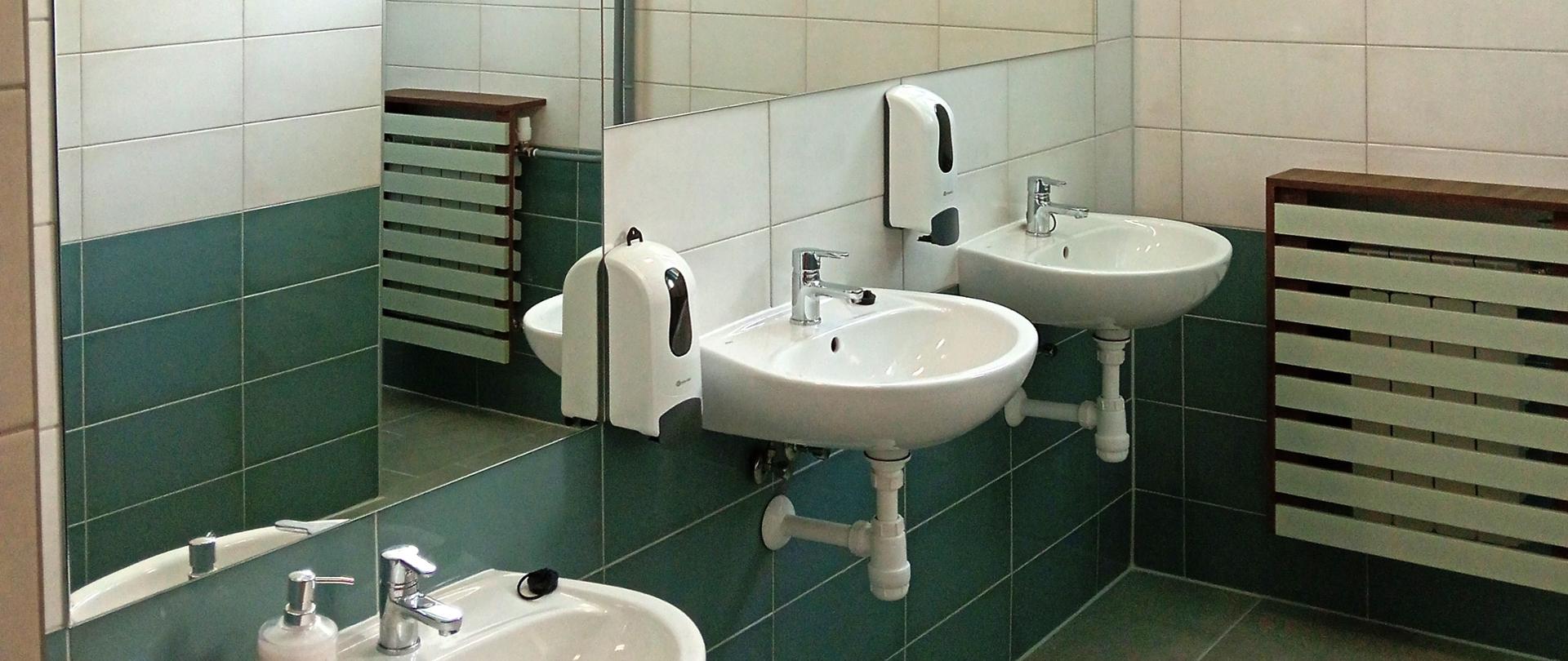 Wnętrze wyremontowanej łazienki z trzema umywalkami, lustrami nad nimi, pojemnikami na mydło i ozdobnie obudowanym kaloryferem, płytki są w kolorze ciemnoniebieskim, nad nimi na panelu zamieszczone kontury trzech postaci kota z myszką