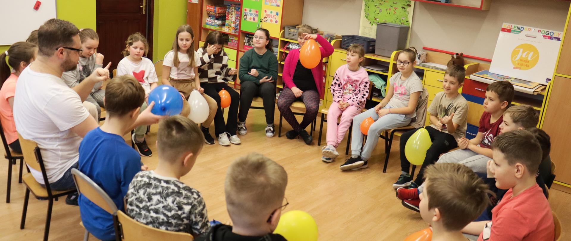 Uczniowie siedzą na krzesłach w kręgu w klasie, niektórzy trzymają w dłoniach balon