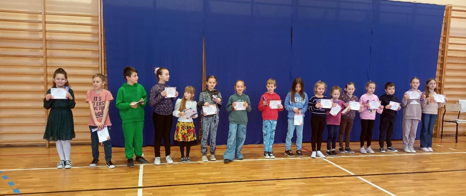 16 dzieci - uczestników konkursu z klas 1-3 stoi na sali gimnastycznej, trzymają w rękach dyplomy zdobyte w konkursie