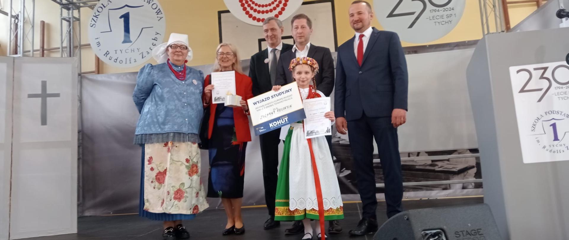 Dziewczynka w ludowym stroju stoi na scenie z kilkoma dorosłymi osobami trzymając dyplomy.