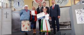 Dziewczynka w ludowym stroju stoi na scenie z kilkoma dorosłymi osobami trzymając dyplomy.