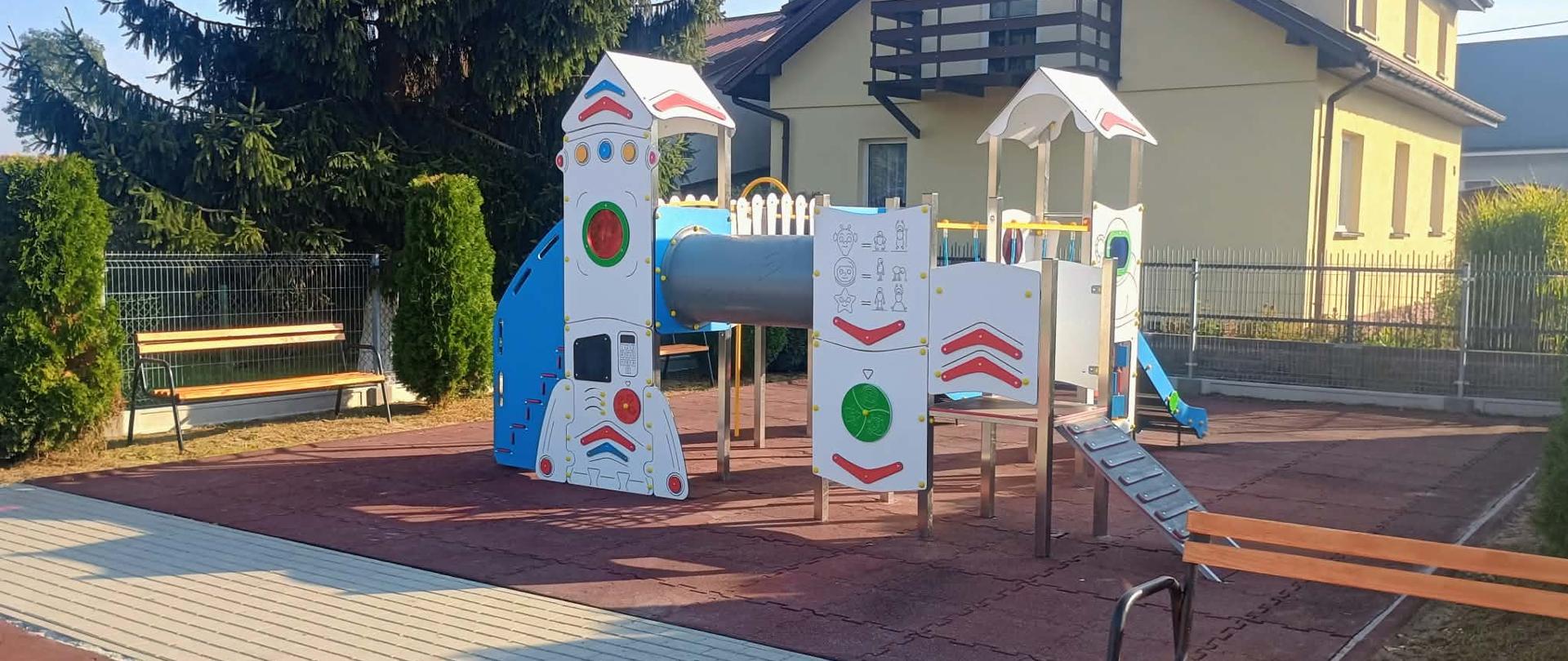 Widok na odnowiony, nowoczesny zestaw zabawowy na placu zabaw w Demblinie. Konstrukcja zadaszona, w kolorach niebieskim, białym, czerwonym i zielonym, ma kształt nawiązujący do rakiety kosmicznej. Posiada tunel czołgowy, tablice edukacyjne, małą zjeżdżalnię i platformy. Całość stoi na bezpiecznej, miękkiej nawierzchni w kolorze ceglanym. Obok widać chodnik z kostki, ławki i wysoki świerk w tle, z domem mieszkalnym na drugim planie.