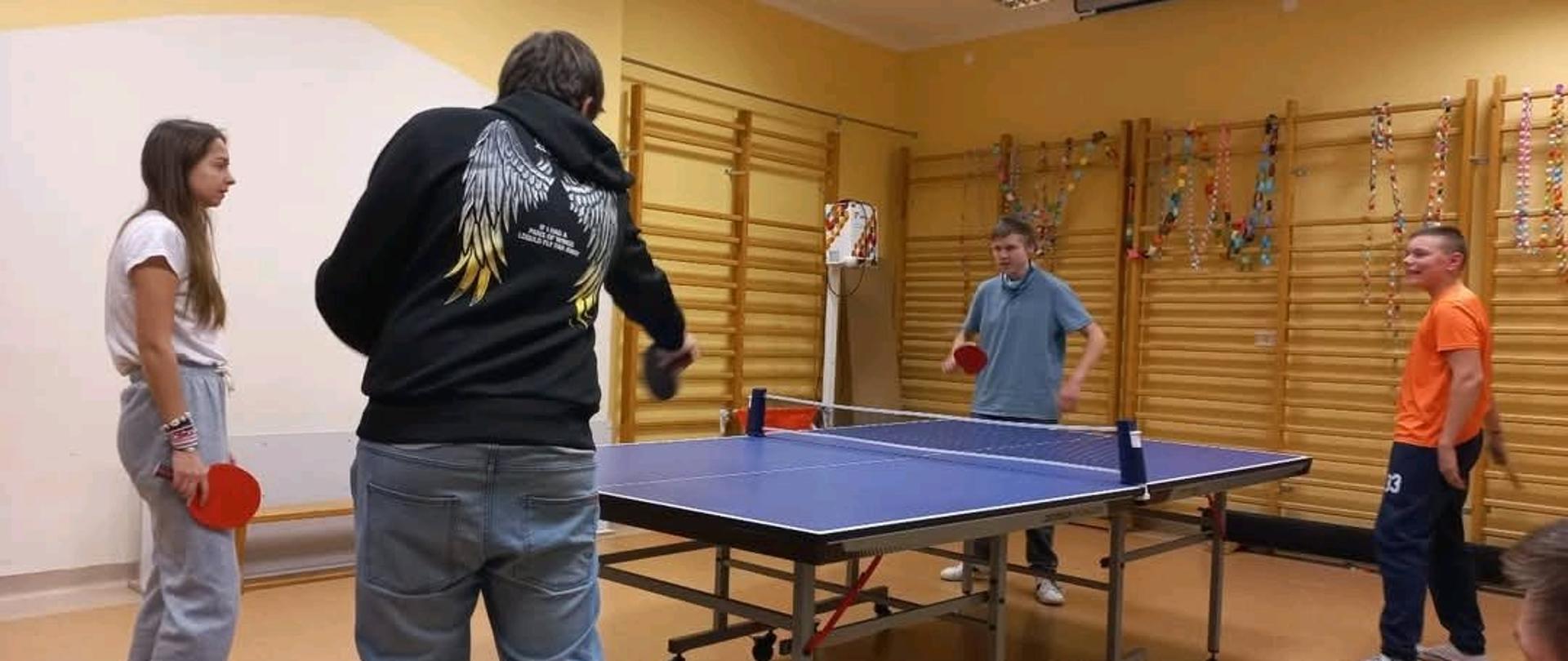 Stół do tenisa stołowego, wokół stołu stoją cztery osoby-trzech chłopców i jedna dziewczyna, w rękach trzymają paletki i grają mecz. Na ścianach widać drabinki gimnastyczne.